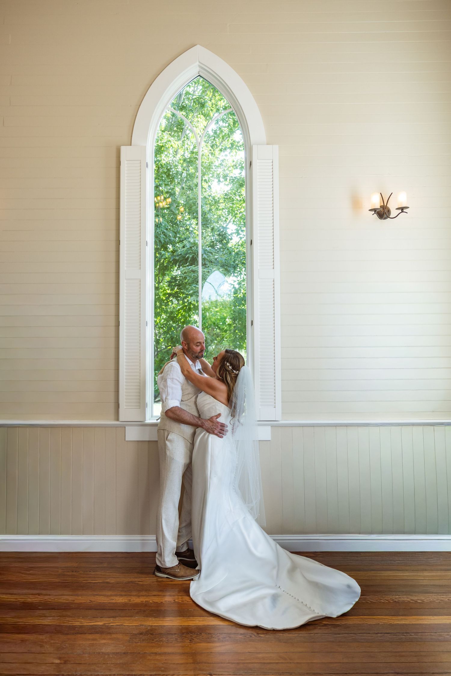 Bride and groom embracing in front of arched chapel window, soft natural light, hardwood floors, elegant white wedding gown.