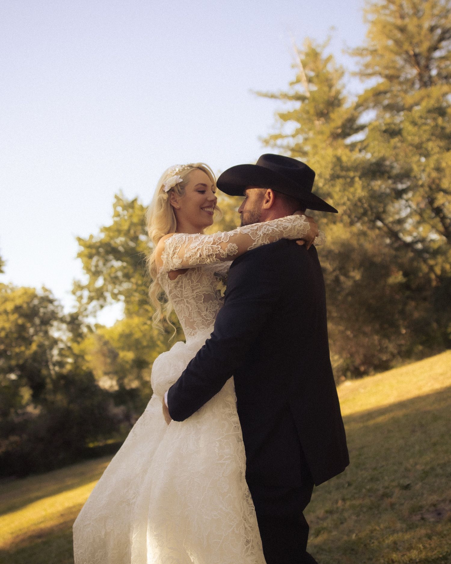 Bride in lace gown and groom in black suit with cowboy hat share romantic dance outdoors at golden hour wedding.