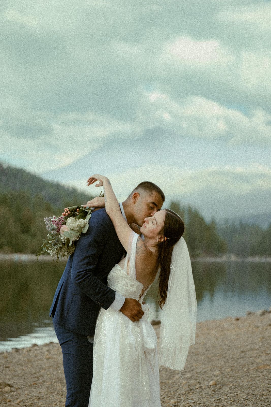 Newlyweds share romantic moments by mountain lake in elegant wedding attire.