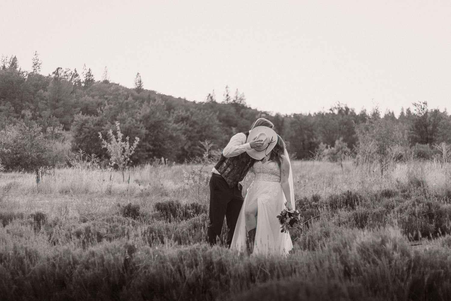 A black and white portrait of a couple embracing in an outdoor field with hills in the background.