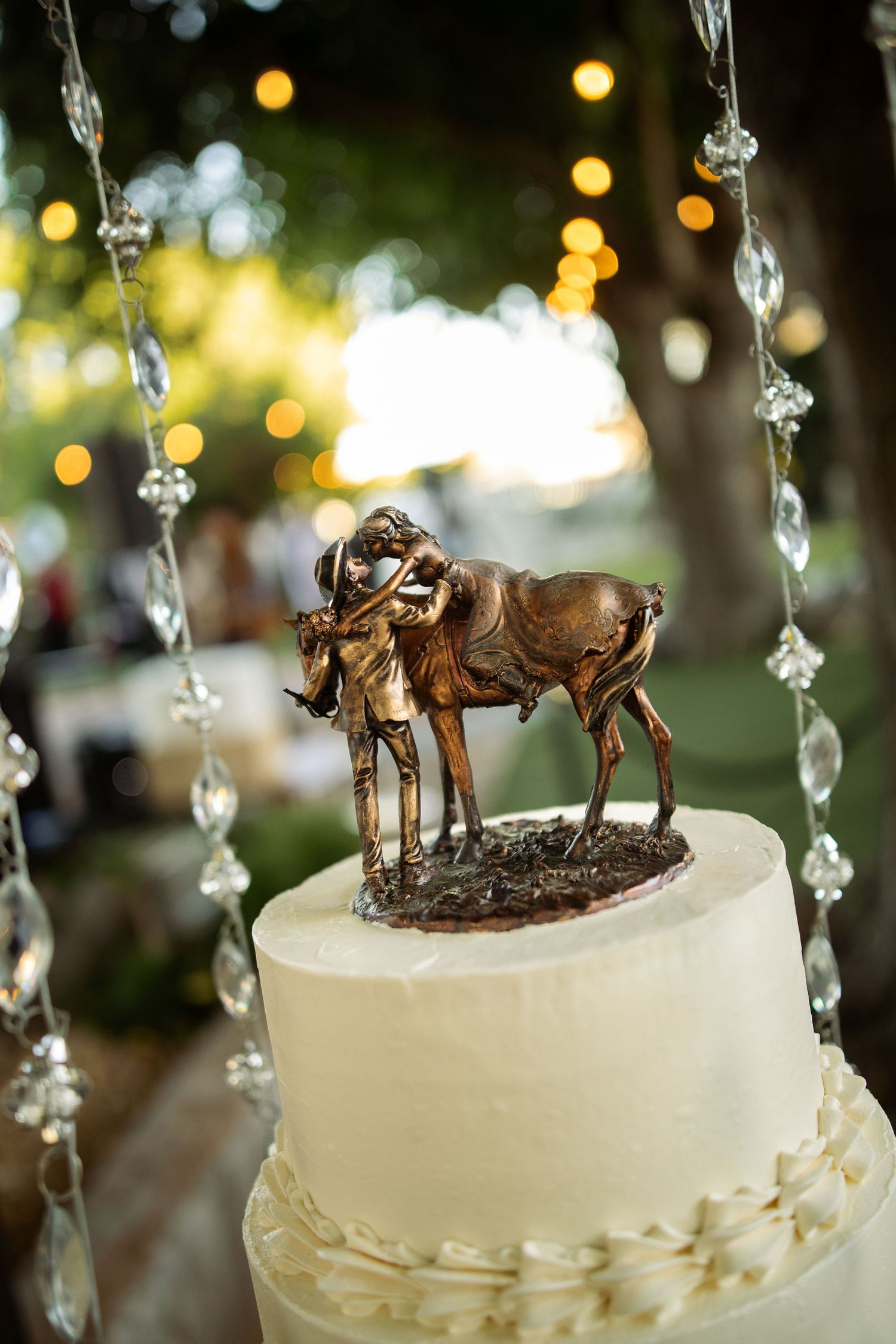 White wedding cake topped with a bronze cowboy and horse figurine topper, surrounded by crystal bead garlands outdoors.