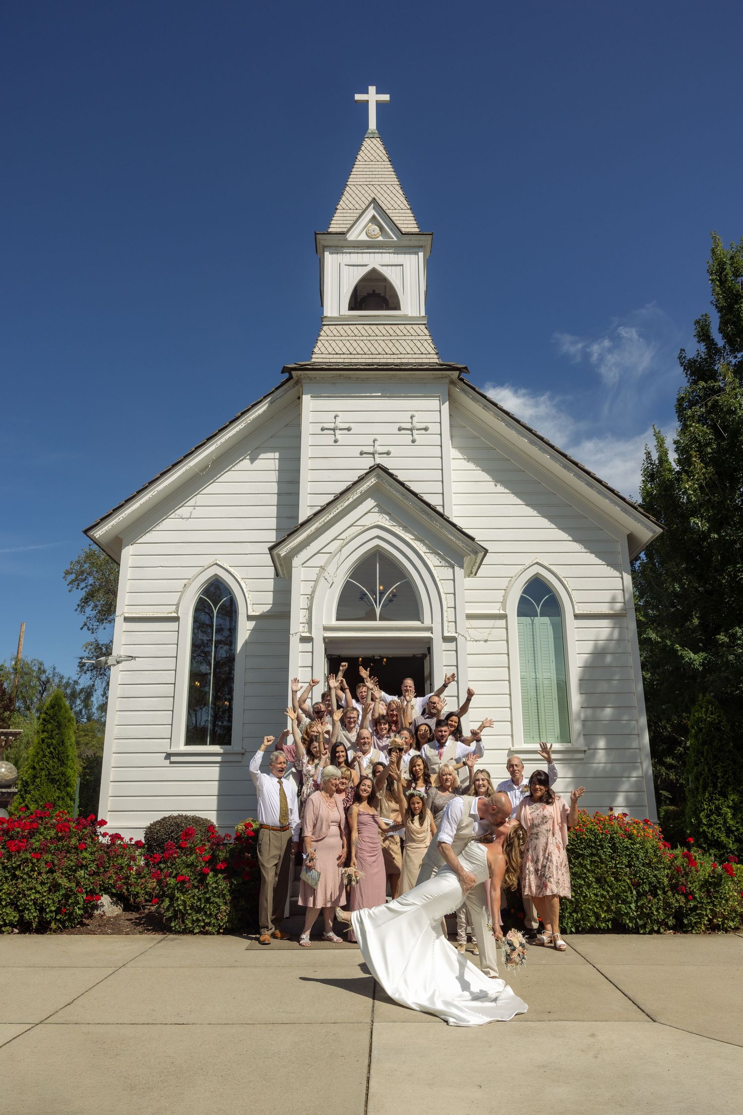Wedding party celebrating outside a white chapel with a cross steeple, bride and groom kissing in front.