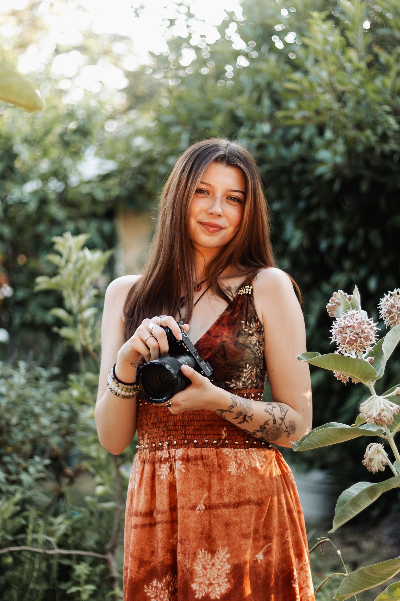 A person in a rust-colored velvet dress poses outdoors among green foliage in a garden setting.