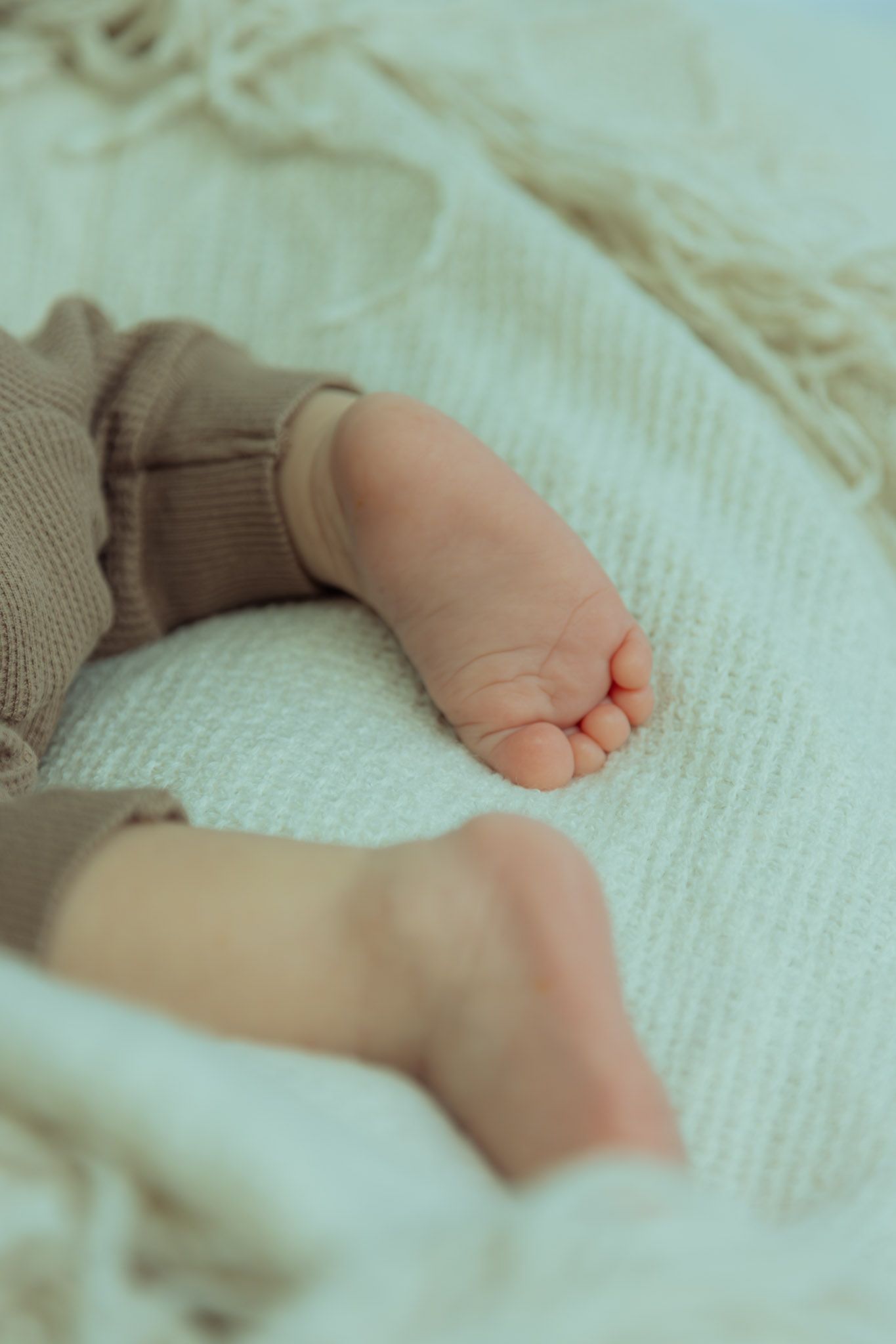 Tiny baby feet with bare toes resting on soft white textured fabric with brown knit clothing visible.
