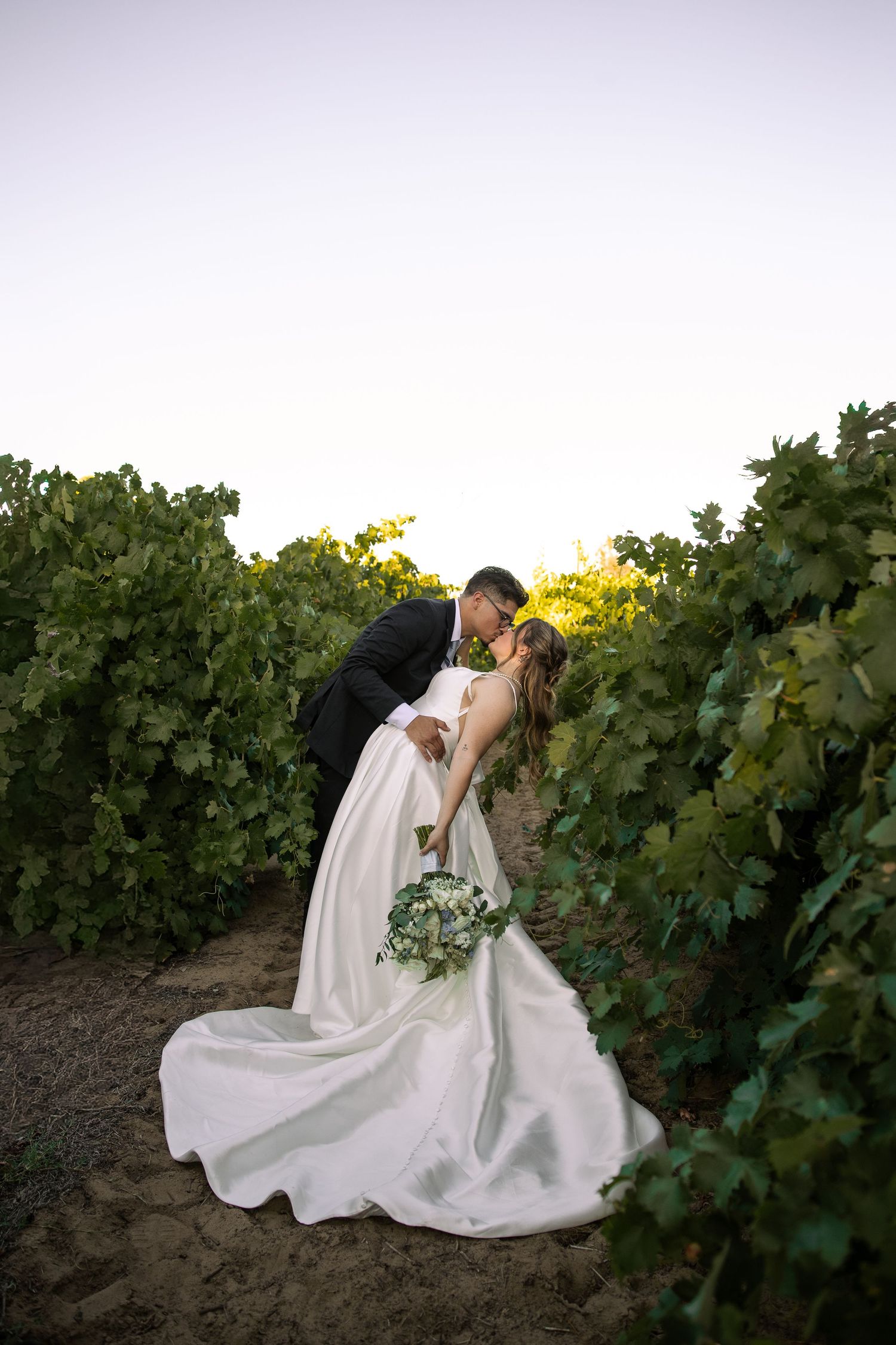 Newlywed couple shares a romantic dip kiss among lush vineyard rows at sunset after their wedding ceremony.