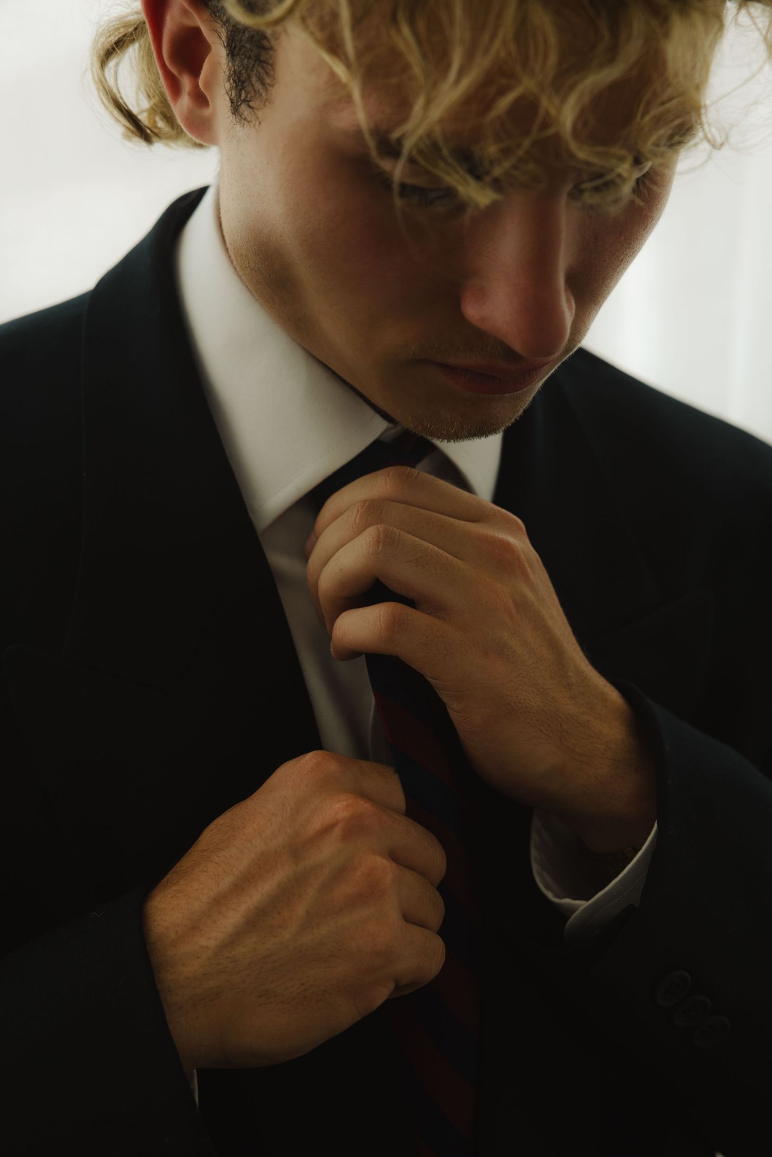 Young man with curly blonde hair adjusting his dark tie while wearing a black suit and white dress shirt.