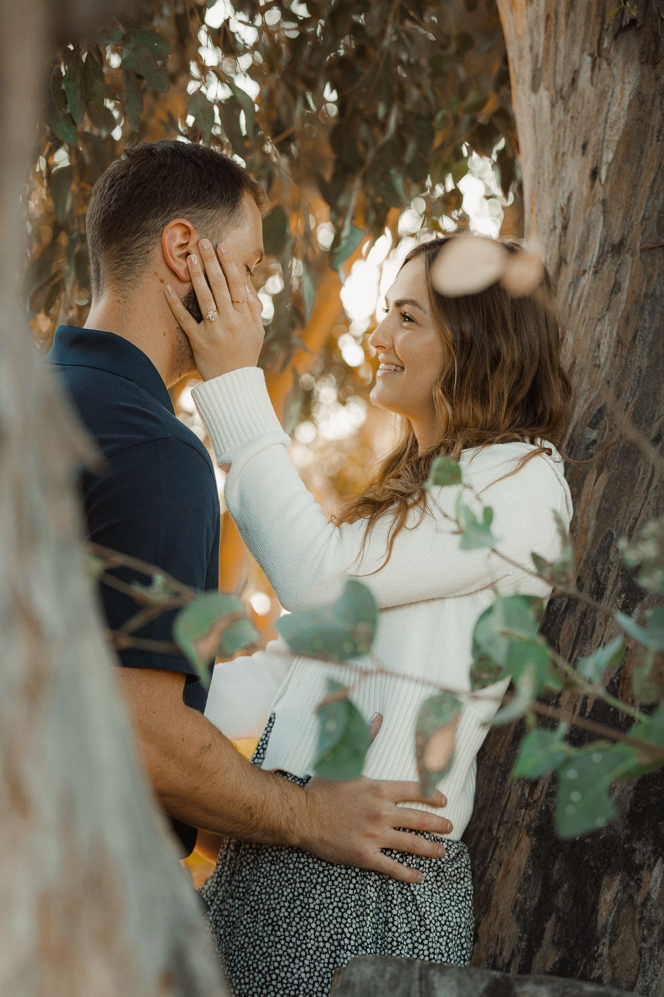 A romantic couple shares an intimate moment among trees at sunset, with soft golden light filtering through the leaves.