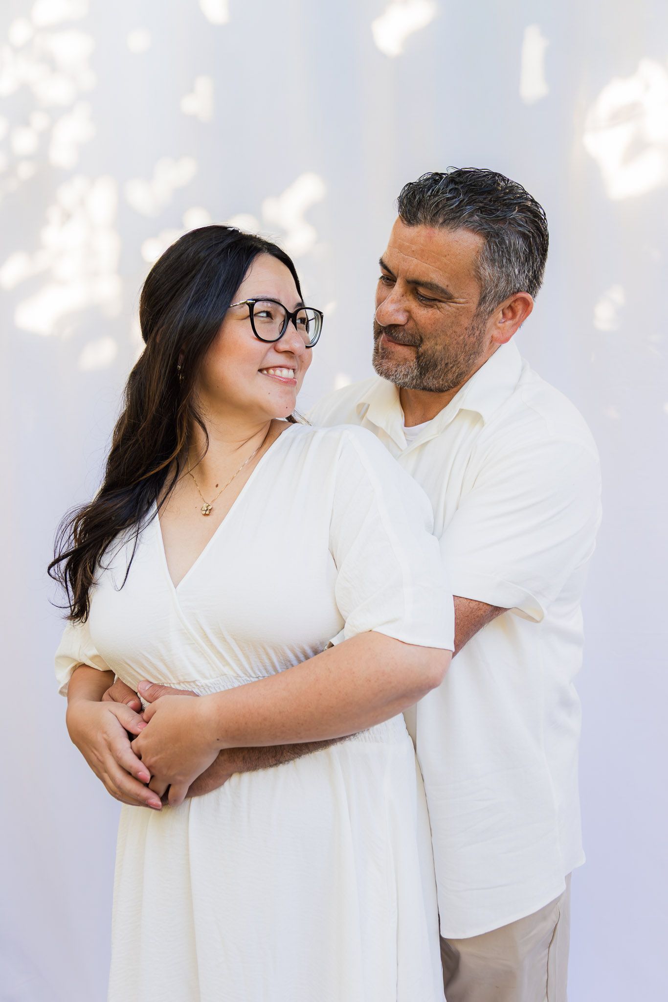 A couple in white outfits embraces lovingly during an outdoor photoshoot with soft lighting and bokeh effects.