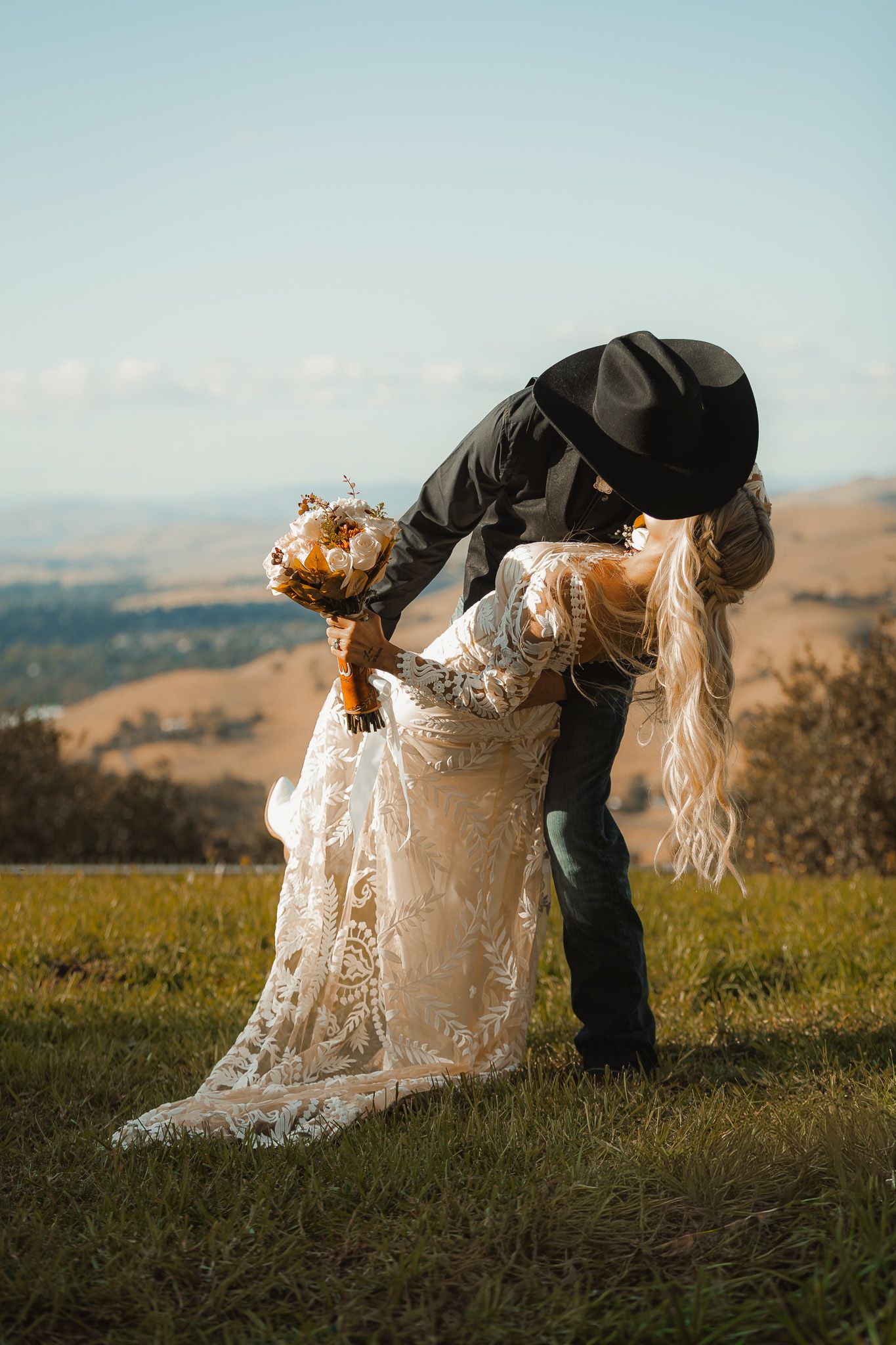 Romantic dip pose of newlyweds on hillside with scenic mountain backdrop.