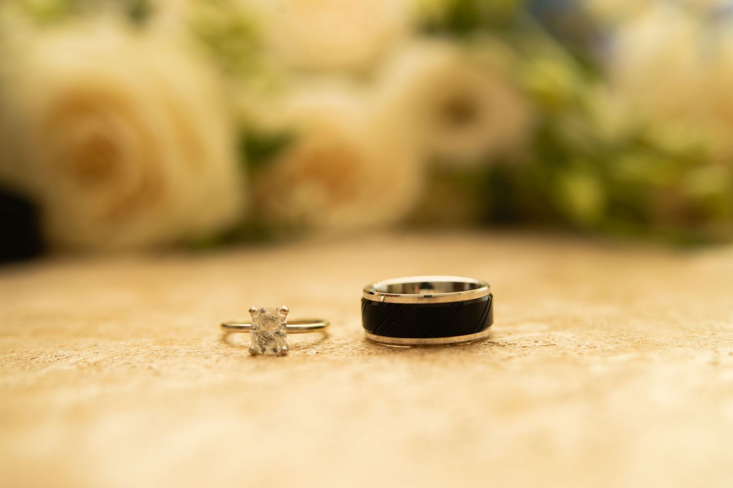 Diamond engagement ring and black tungsten wedding band displayed on a marble surface with white roses in the background.