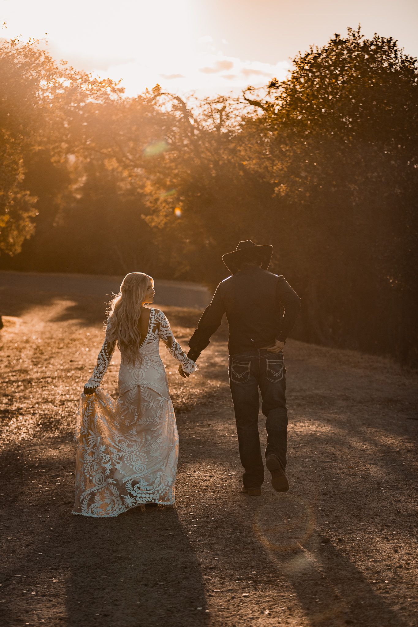 Newlyweds walk hand in hand during golden hour in rustic outdoor setting.