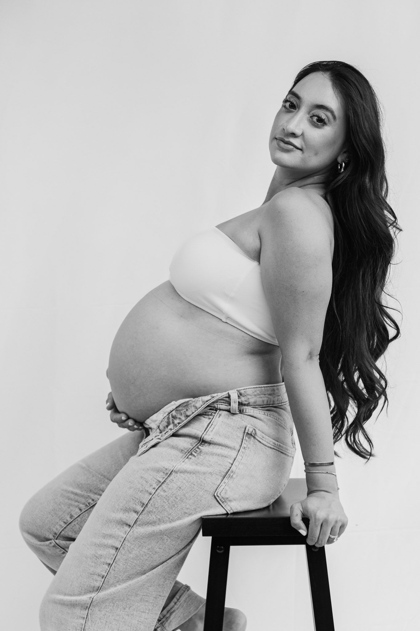 Maternity portrait of pregnant woman in casual attire posing on stool against white background.