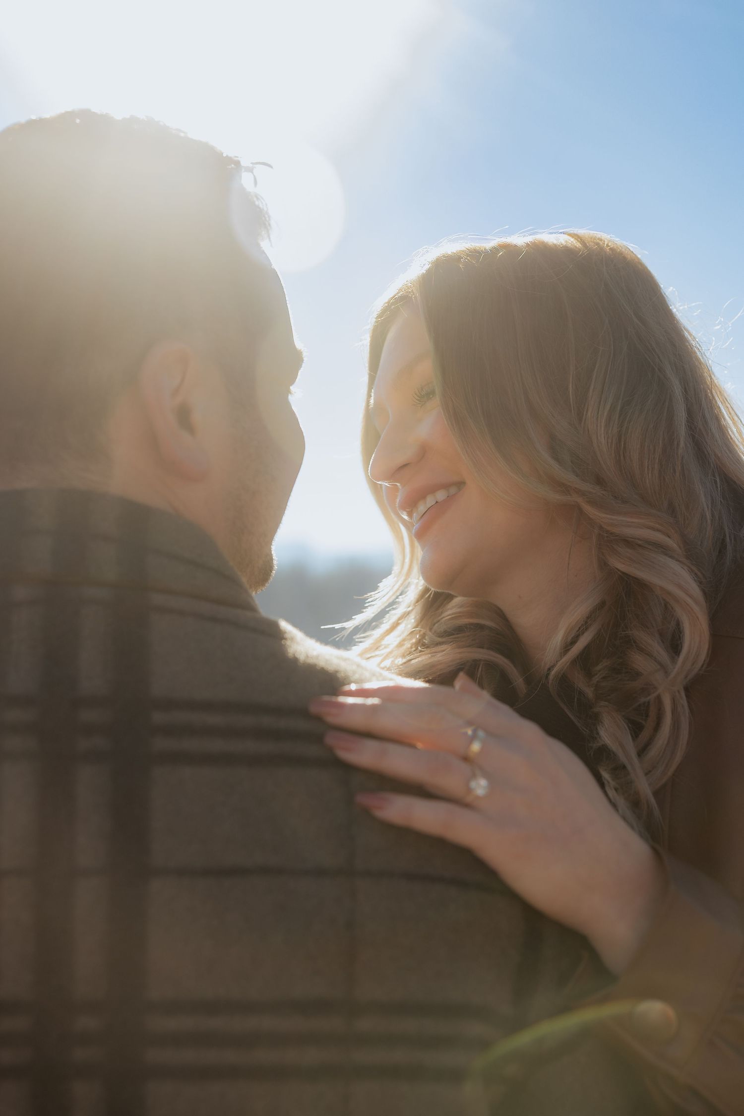 A romantic couple embraces in warm sunlight, with the woman's engagement ring visible as she touches her partner's shoulder.