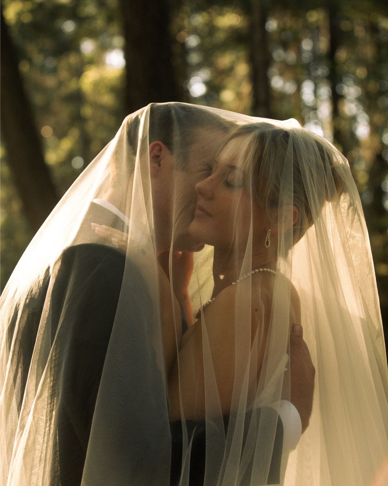 Bride and groom sharing a romantic kiss under a flowing wedding veil in a sunlit forest setting.