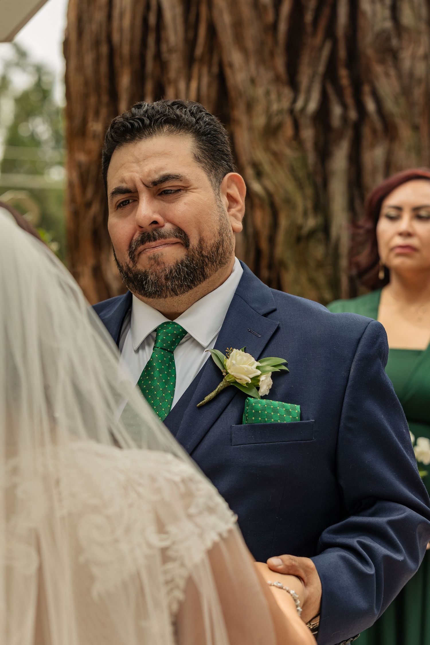 Emotional groom in blue suit with green tie tearing up seeing bride at outdoor wedding ceremony.