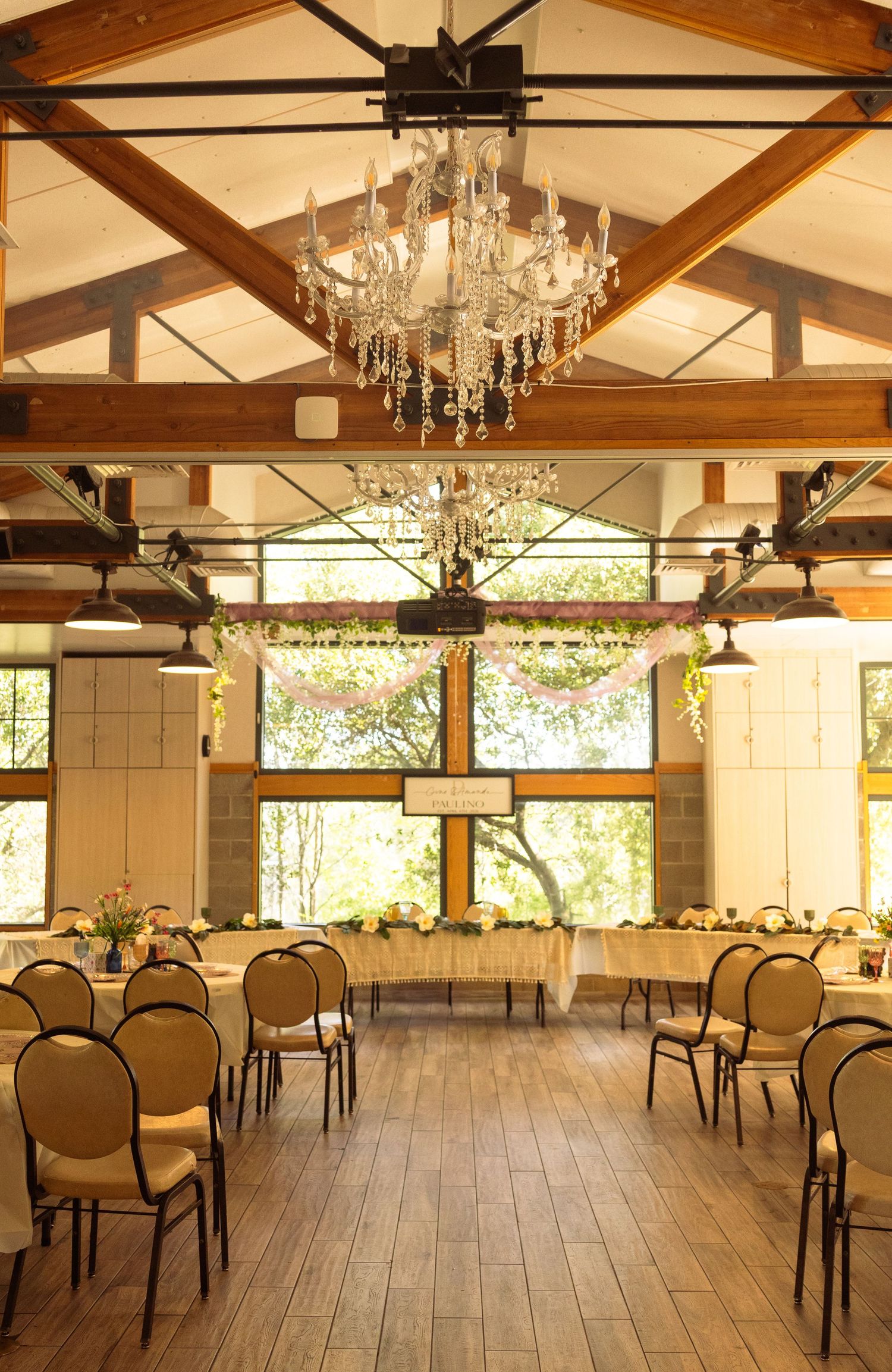 Elegant wedding reception hall with crystal chandelier, wooden beam ceiling, round chairs, and floral decorations by large windows.