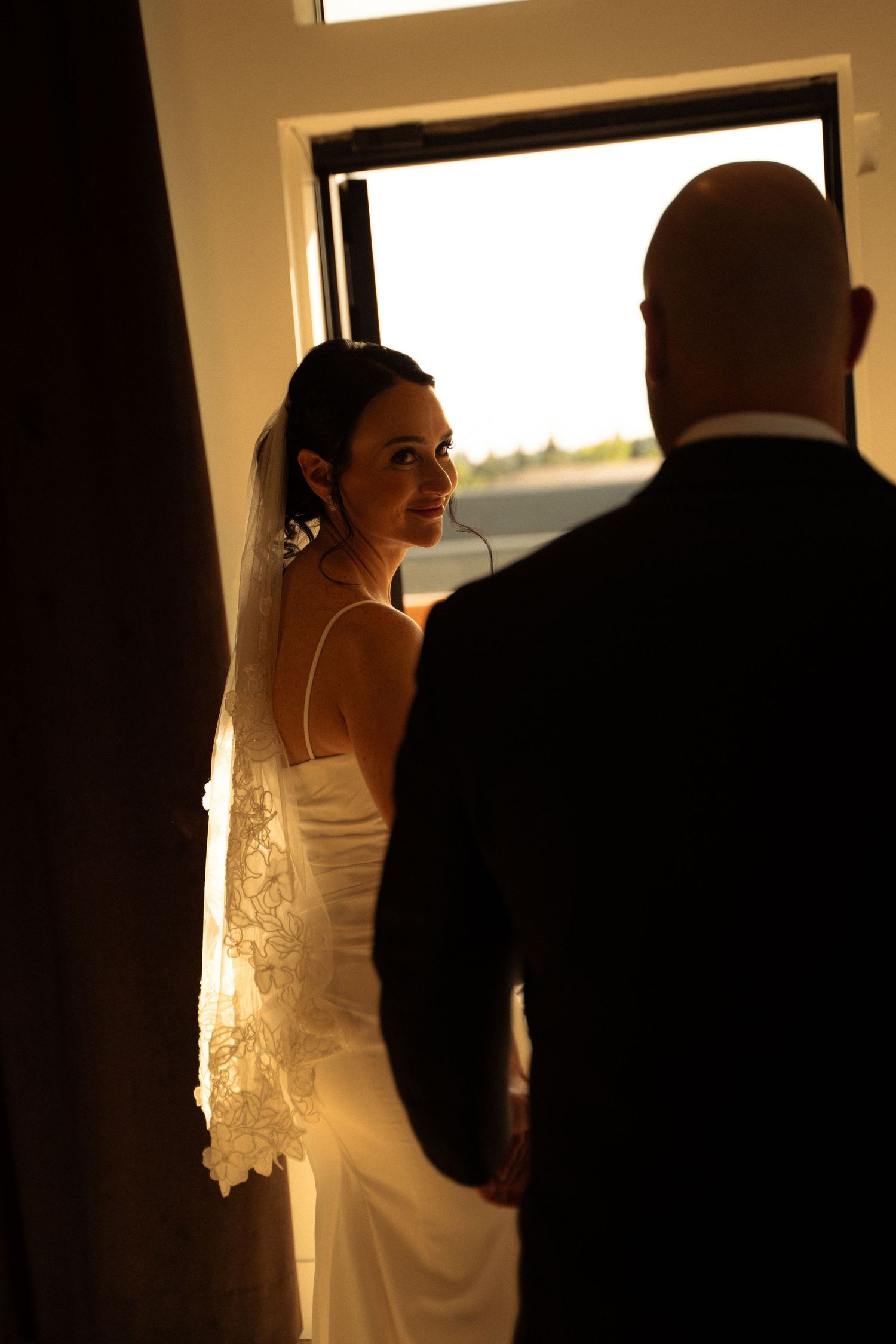 Bride in white gown and veil glances back at bald groom in tuxedo standing by sunlit window.