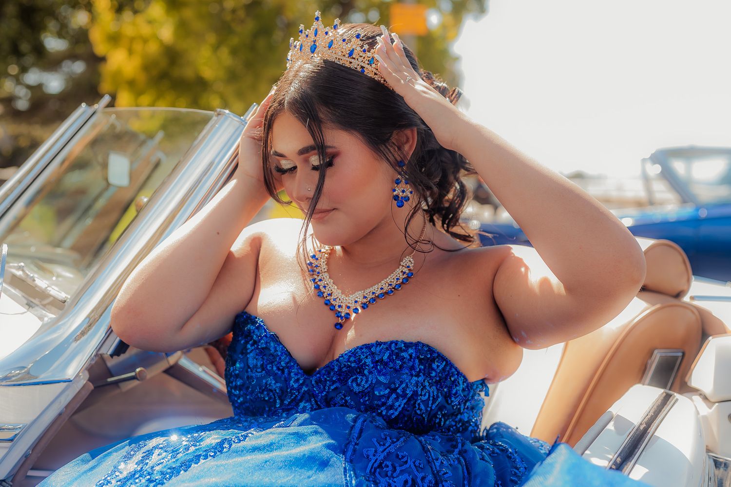 A person in a sparkling blue formal gown poses elegantly near a classic car on a sunny day.
