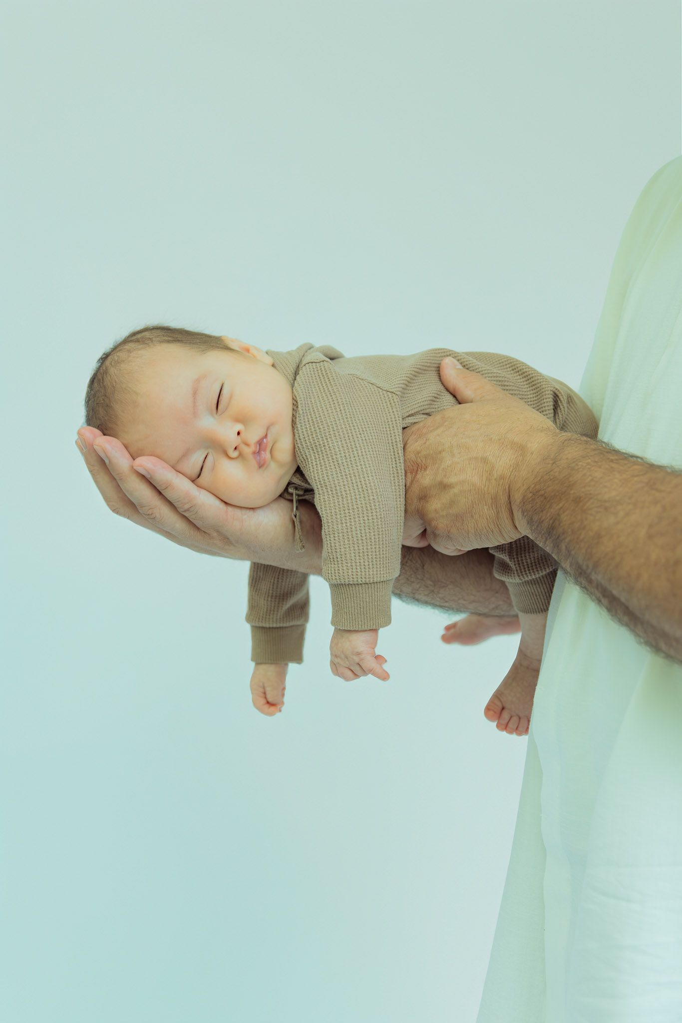 A sleeping baby wearing a tan onesie is gently cradled in adult hands against a mint green background.