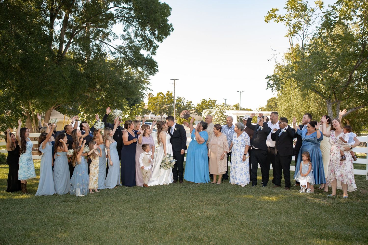 Large wedding party and guests pose together on a green lawn surrounded by trees during an outdoor celebration.