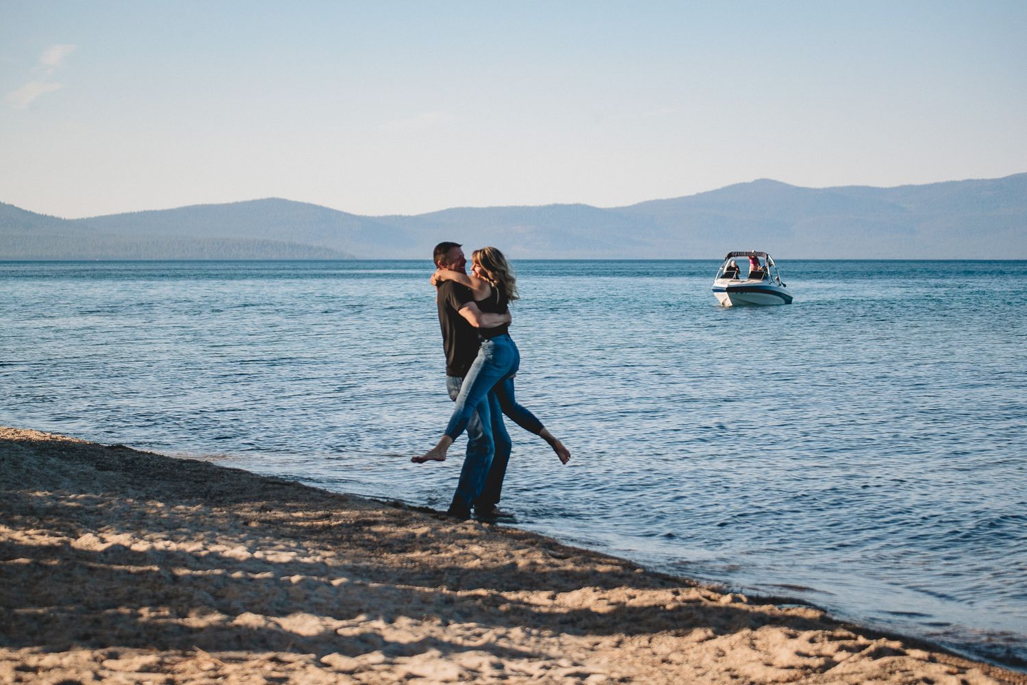 A couple embraces on a scenic beach with mountains and a boat in the distance on a clear sunny day.