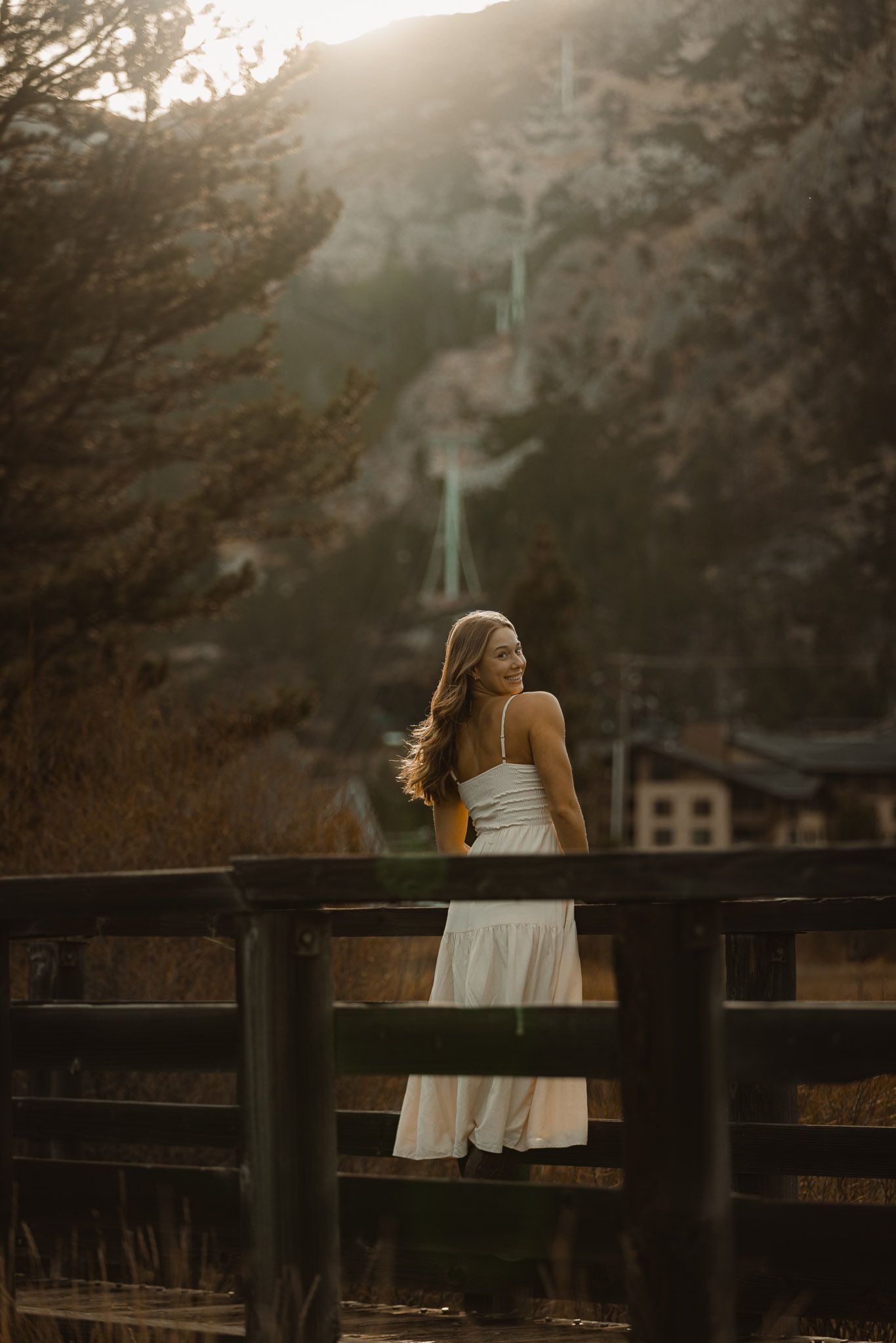 Person in white dress leaning on wooden fence at sunset with mountains in background.