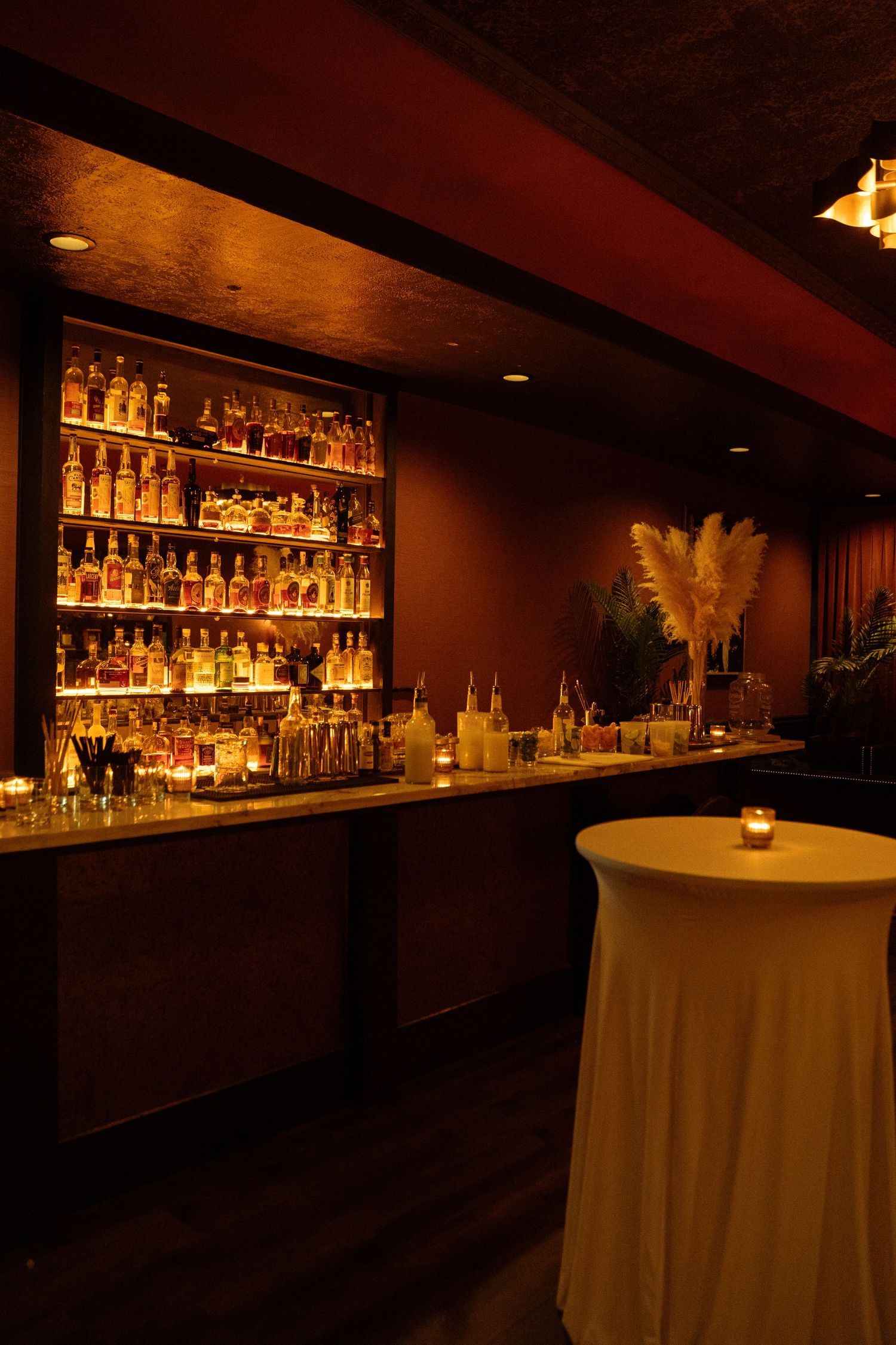 Dimly lit upscale bar with backlit liquor shelves, marble counter, and elegant white cocktail table with pampas grass.