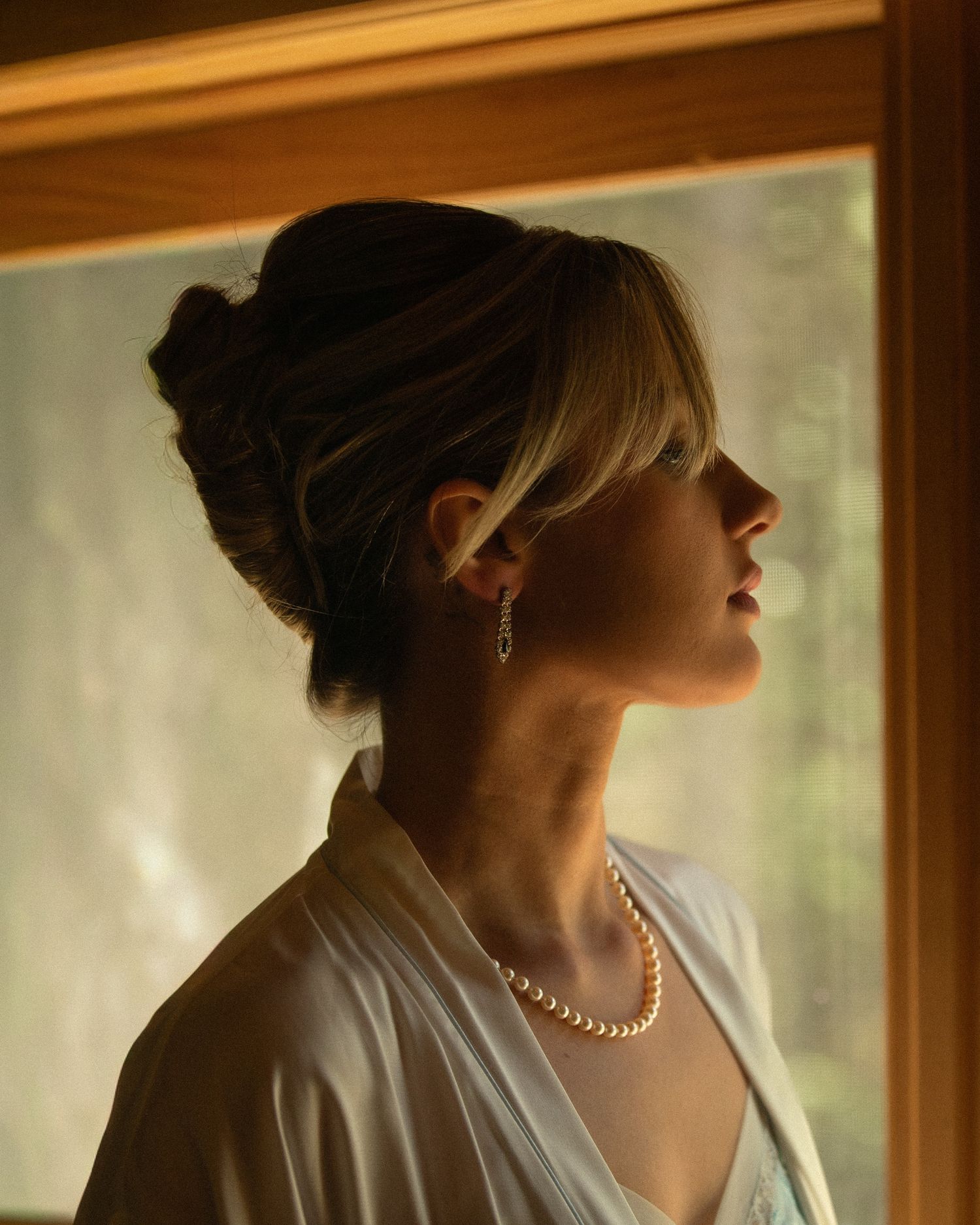 A woman in a white robe with pearl necklace and earrings gazes upward in soft warm light.