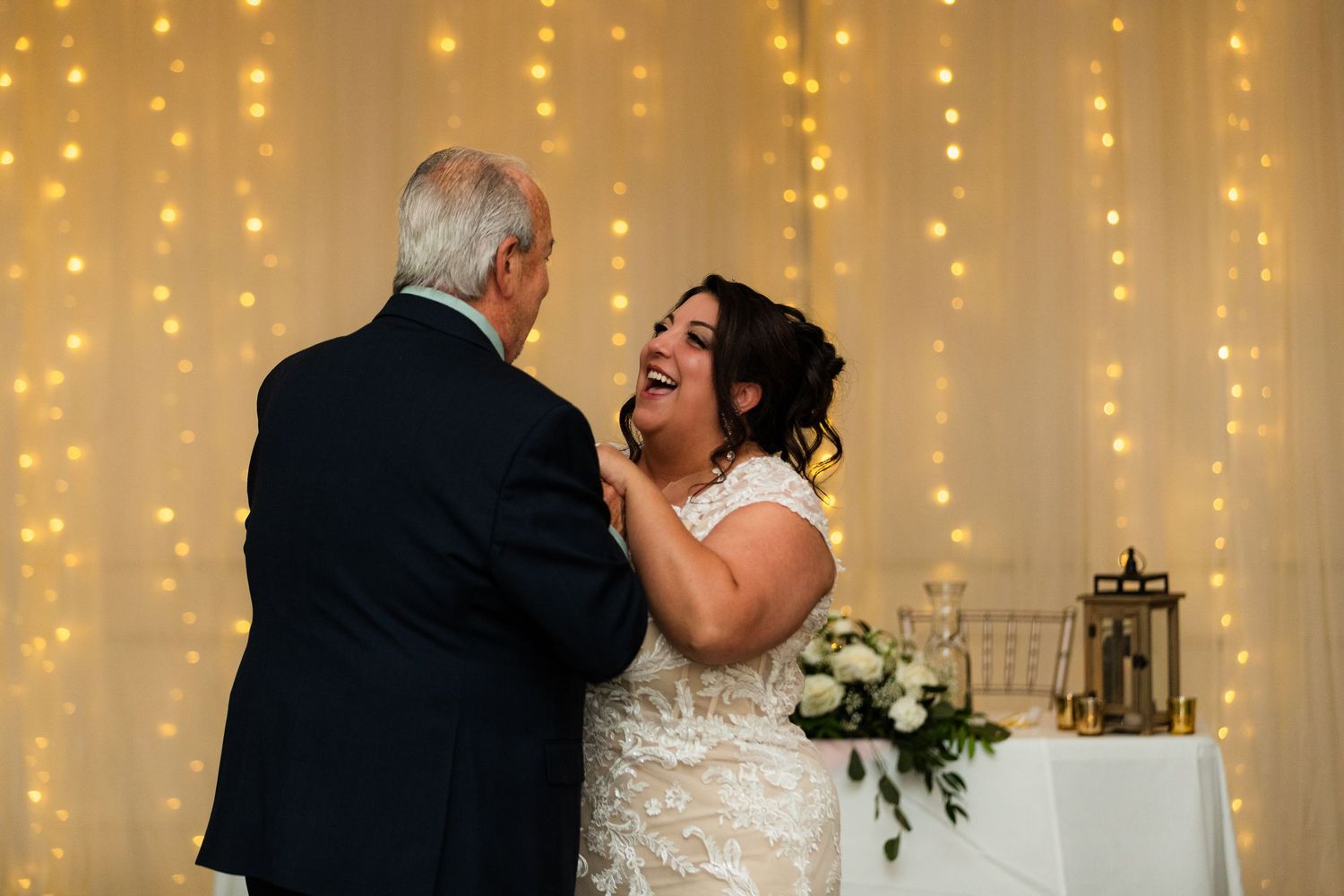 Bride laughing joyfully with older man during father-daughter dance at elegant wedding reception with fairy light backdrop.