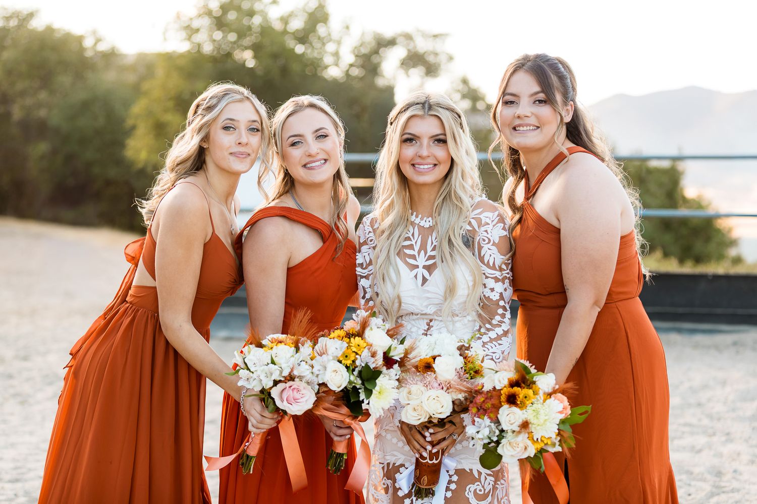Bridesmaids in matching burnt orange dresses pose with bride in lace gown holding autumn bouquets.