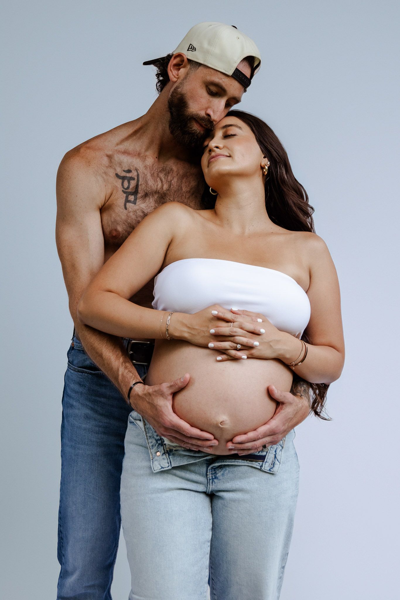 Intimate maternity photography session featuring couple in casual denim and white against grey backdrop.