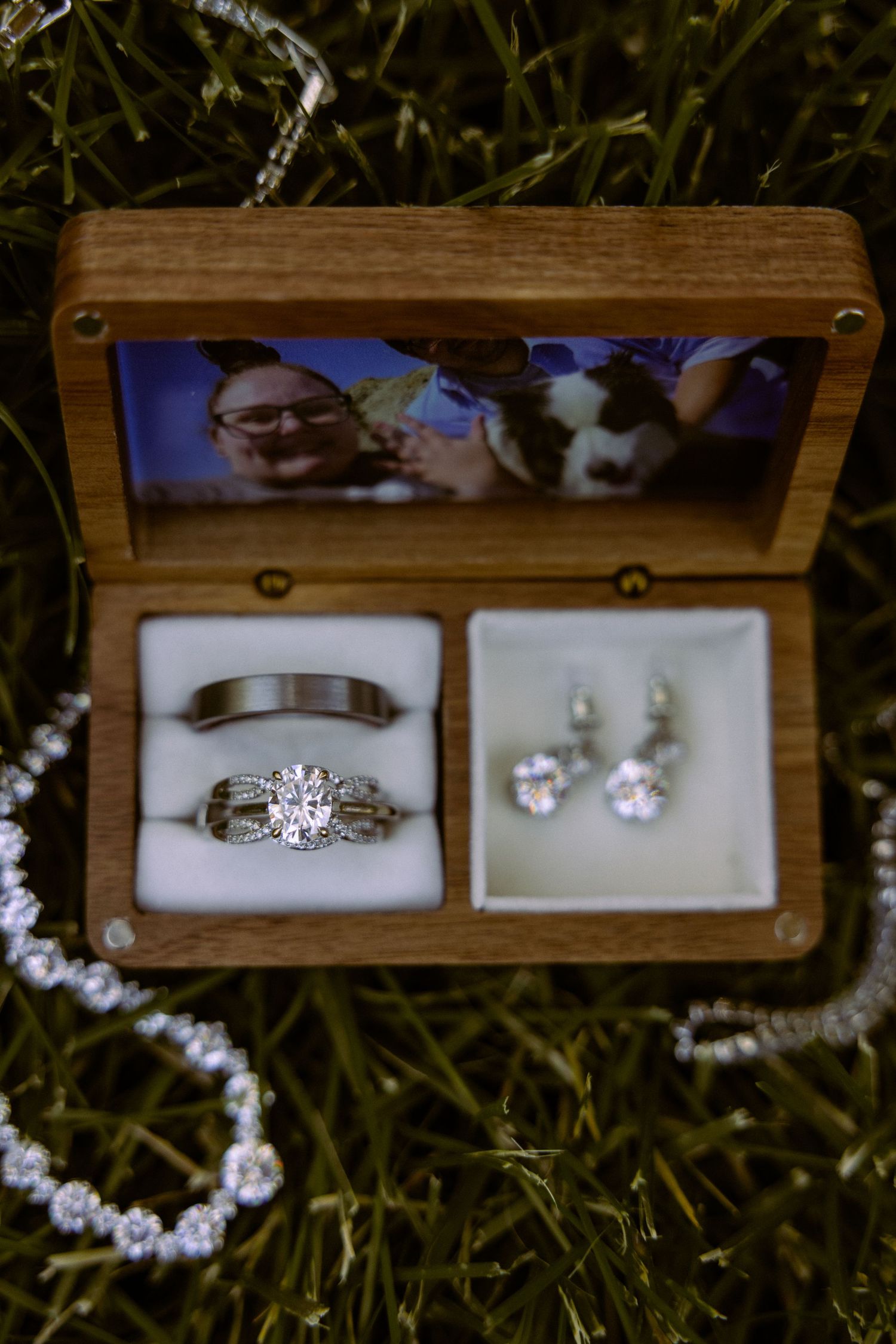Wooden wedding ring box with diamond engagement ring, band, and earrings on grass, featuring a couple photo inside the lid.
