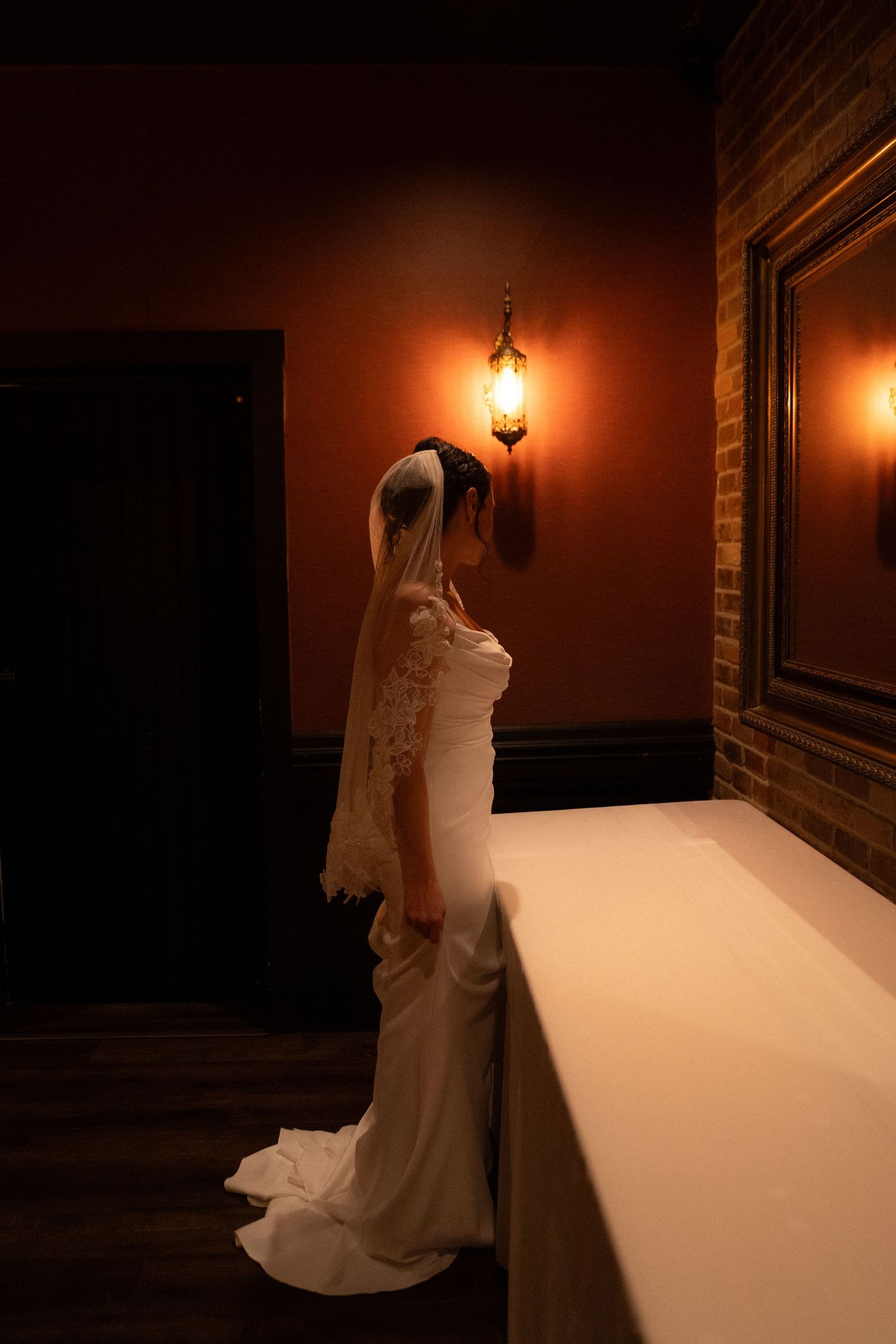 Bride in white gown and veil standing by candlelit wall sconce in dimly lit room.