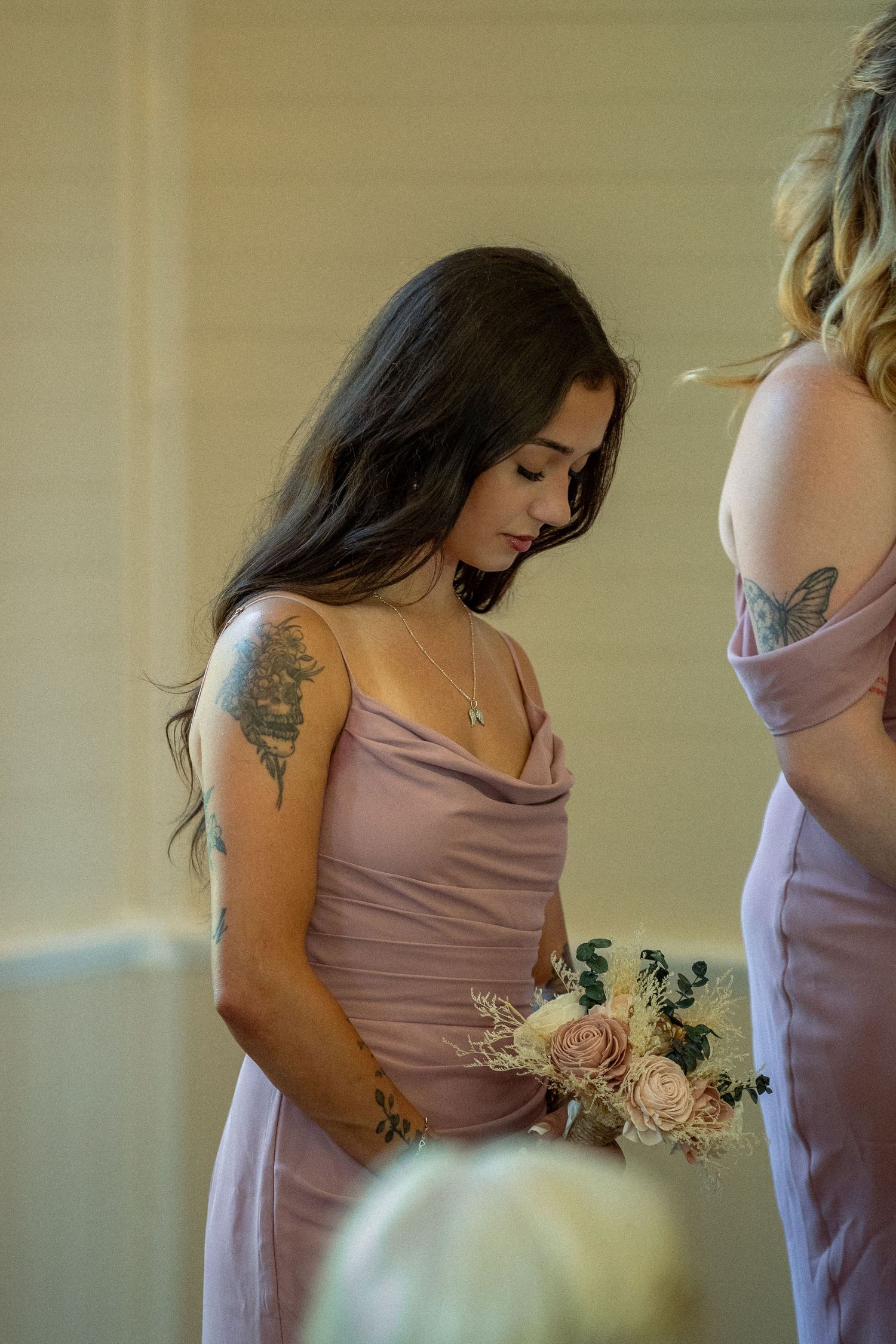 Tattooed bridesmaid in lavender satin gown holding blush floral bouquet, looking down elegantly.