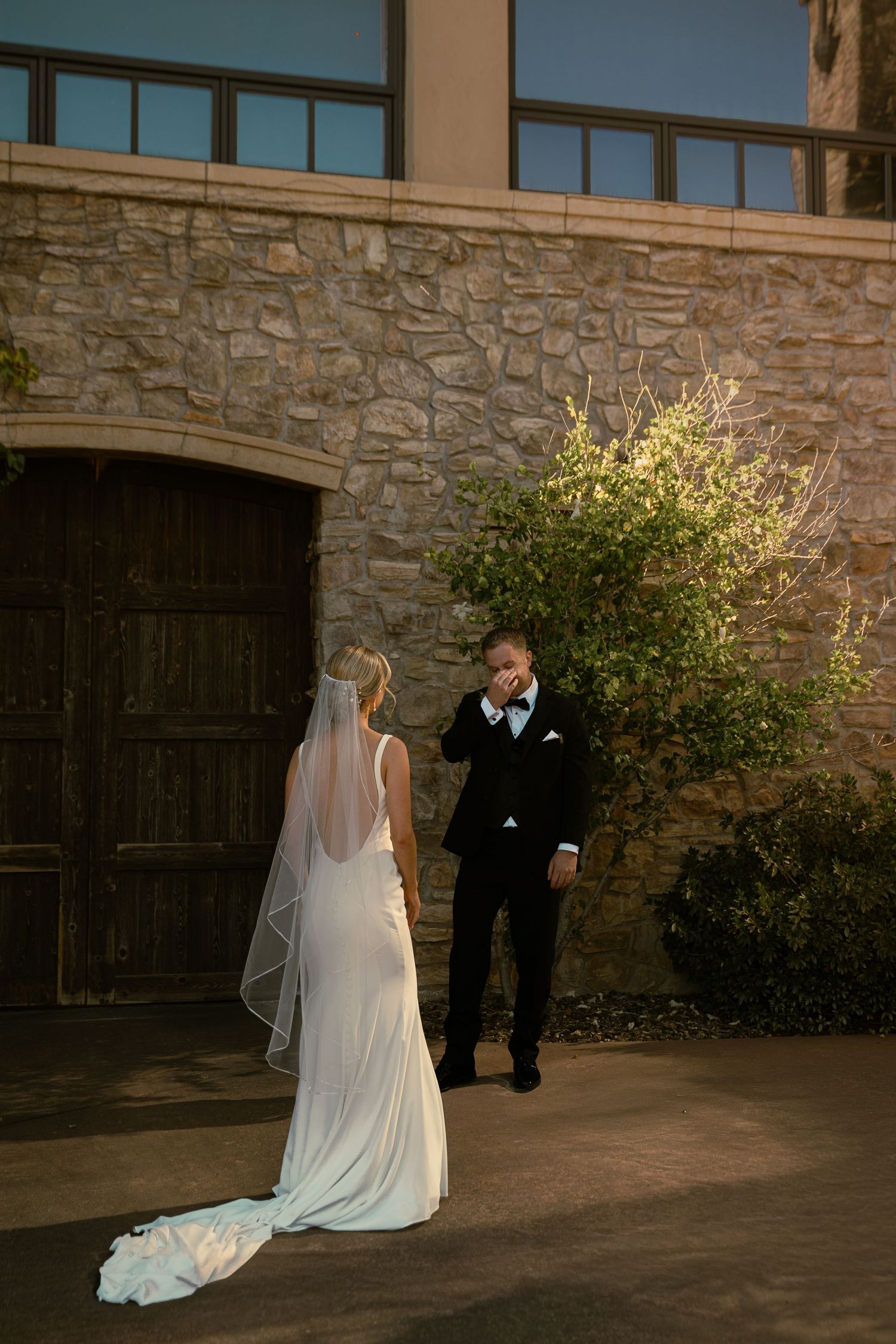 Bride in white gown and veil faces emotional groom in black tuxedo during first look outside stone church.