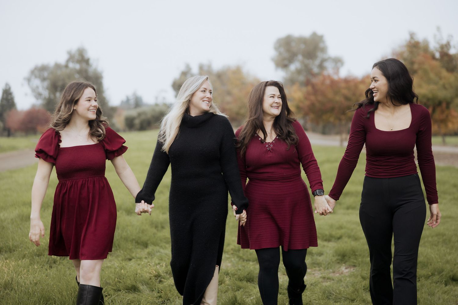 Three friends in burgundy and black outfits walk together laughing in a sunny autumn field.