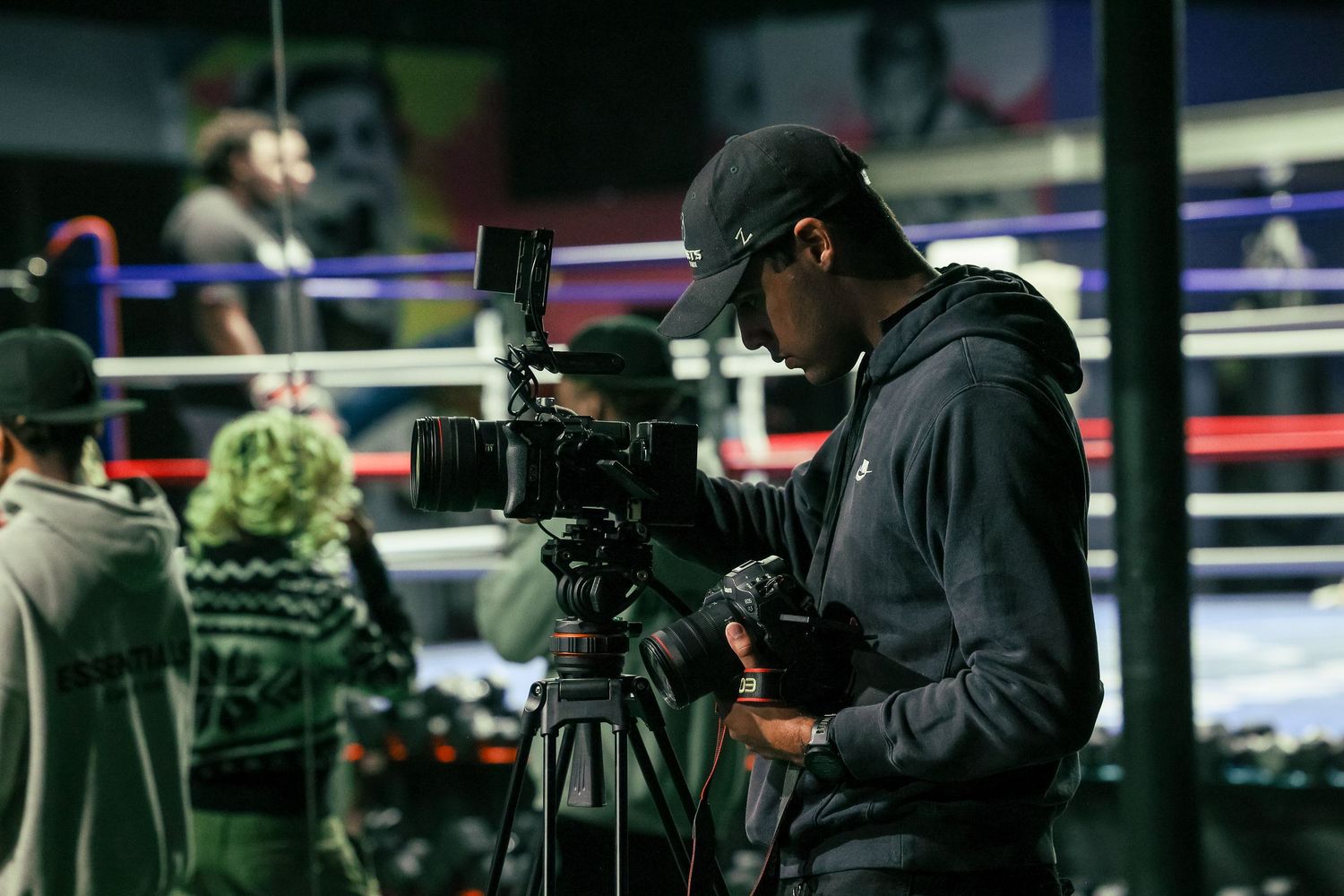 Camera operator films at a boxing ring using professional video equipment while wearing a black hoodie and cap.