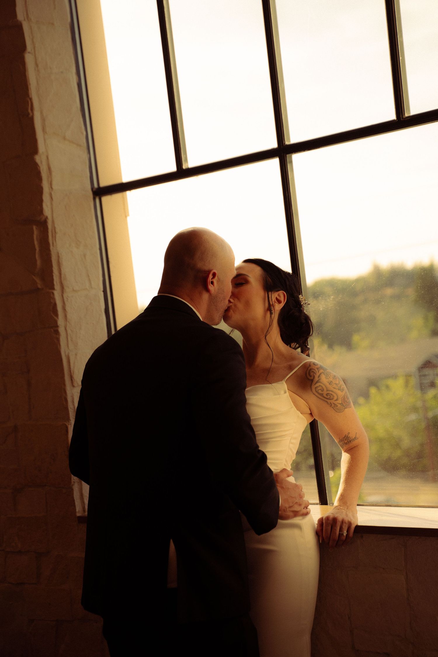 A bride and groom share a romantic kiss silhouetted against a large window on their wedding day.
