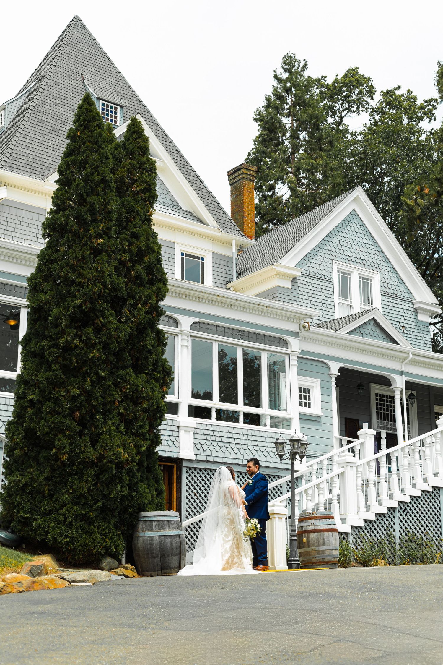 A bride and groom stand before a charming blue Victorian mansion with white trim, flanked by a tall evergreen tree.