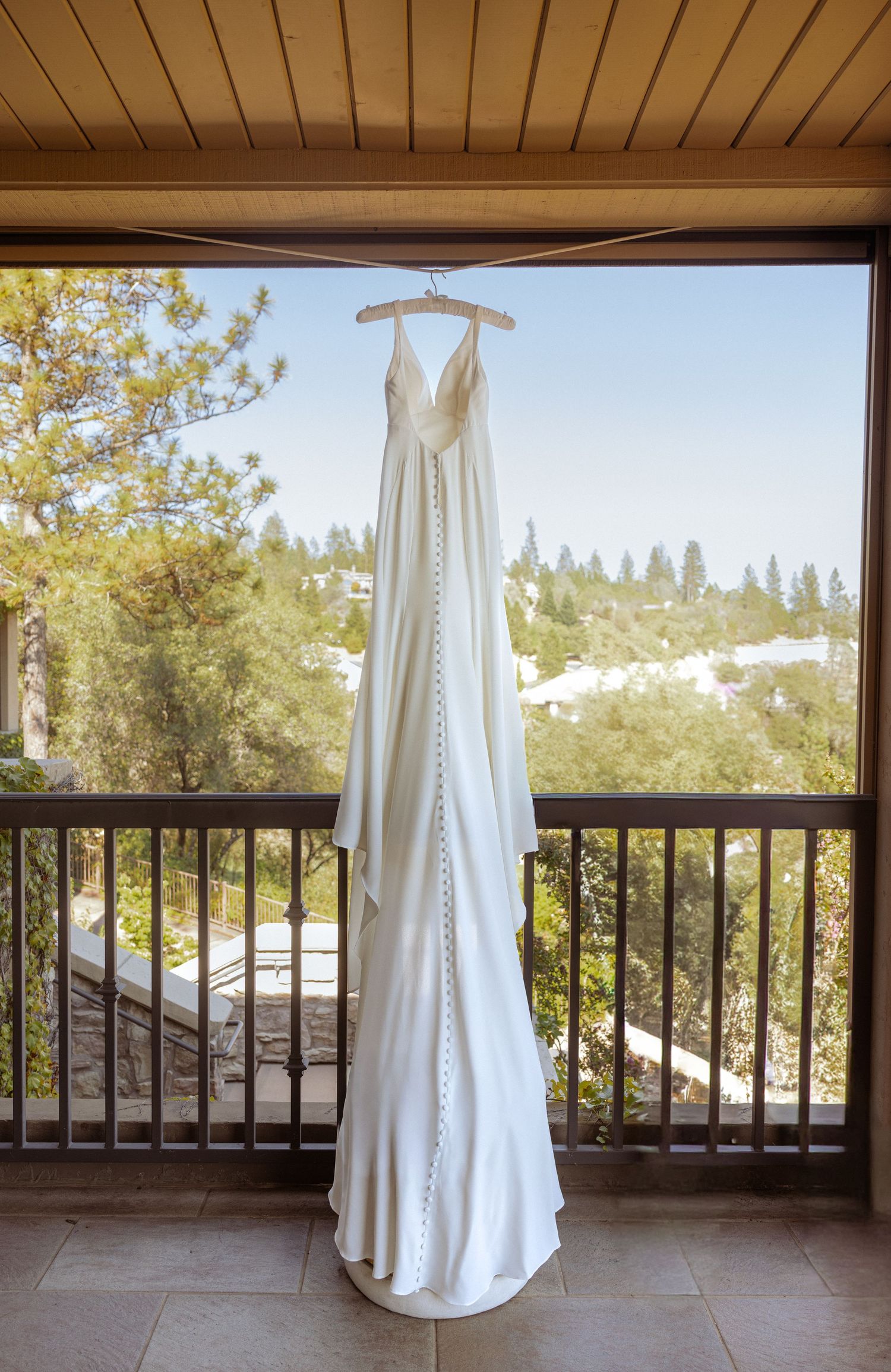Elegant white satin wedding gown with button back detail hanging on balcony overlooking lush green trees.