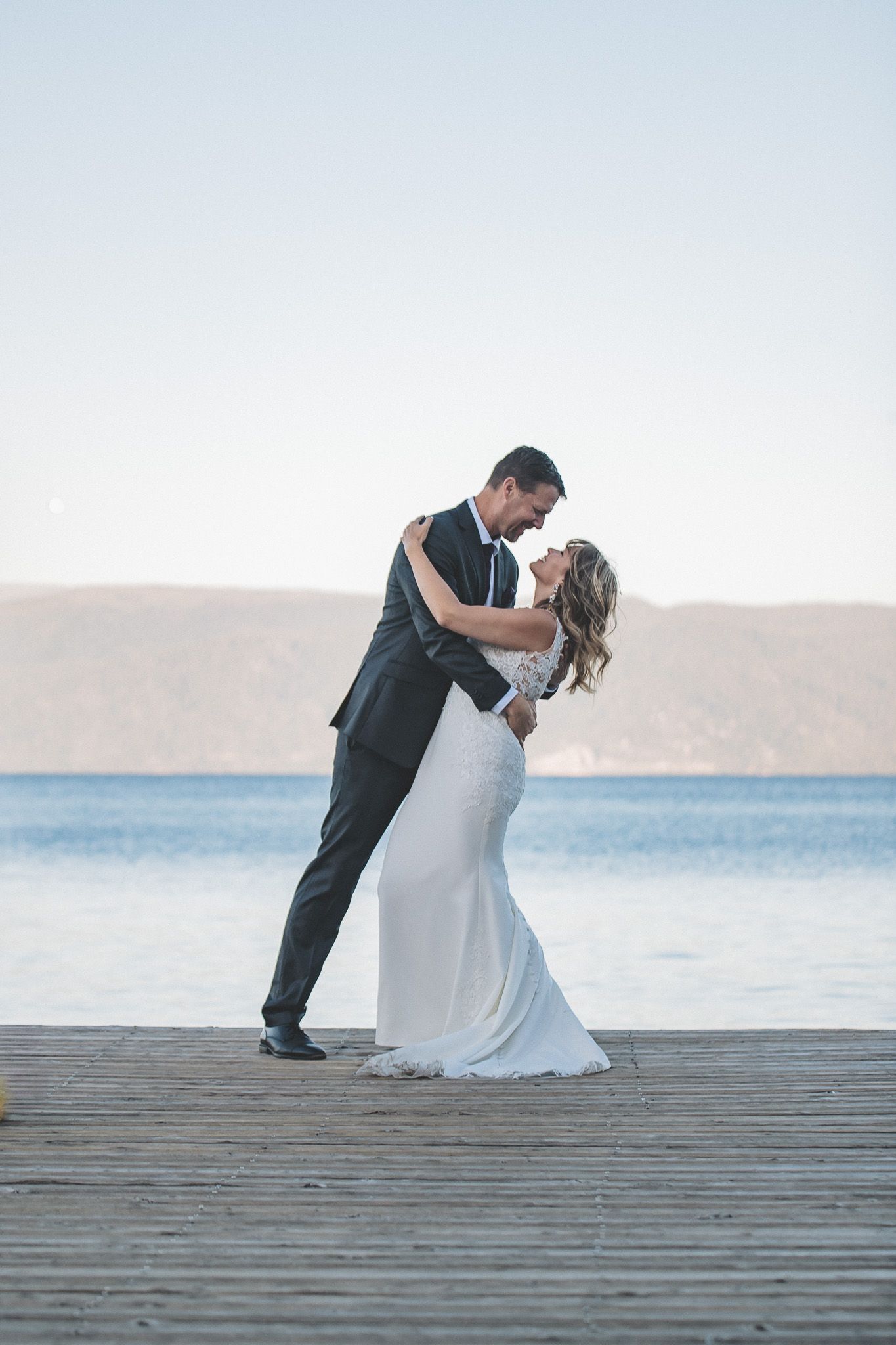 A romantic wedding couple embraces on a wooden dock overlooking a serene lake and mountains at sunset.