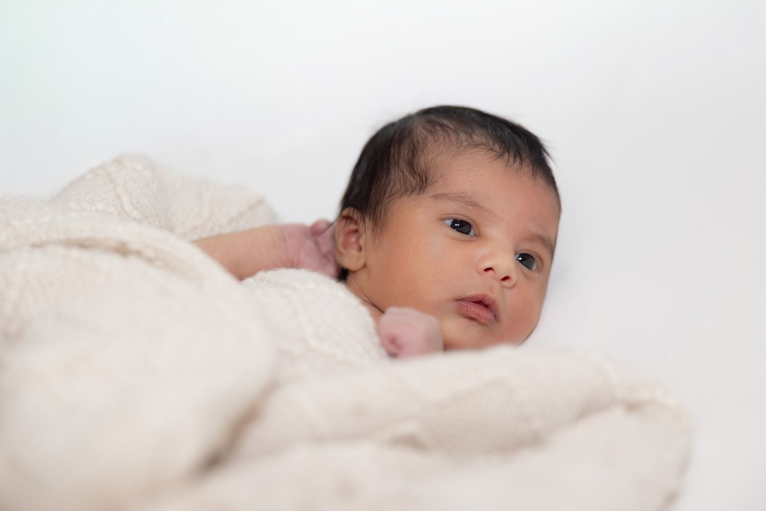 A small baby wrapped in a soft white blanket gazes at the camera with wide eyes.
