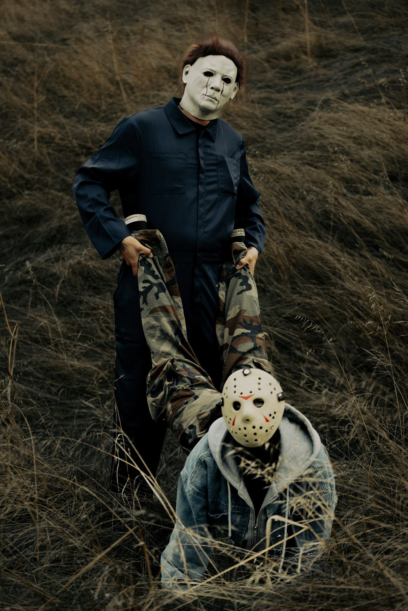 A figure in dark clothing poses with a hockey mask in tall dried grass.