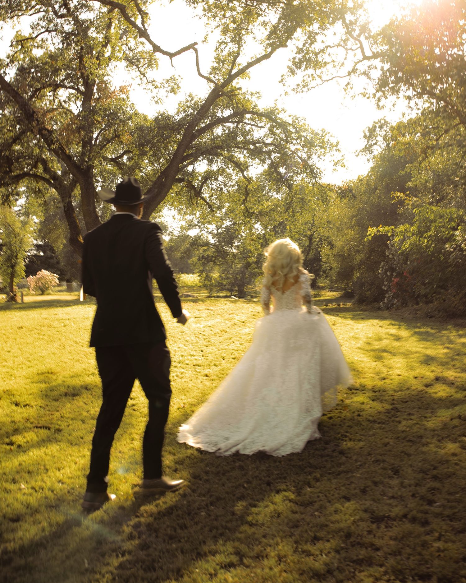 A bride in a white gown and groom in a black suit walk hand in hand through a sunlit green park.