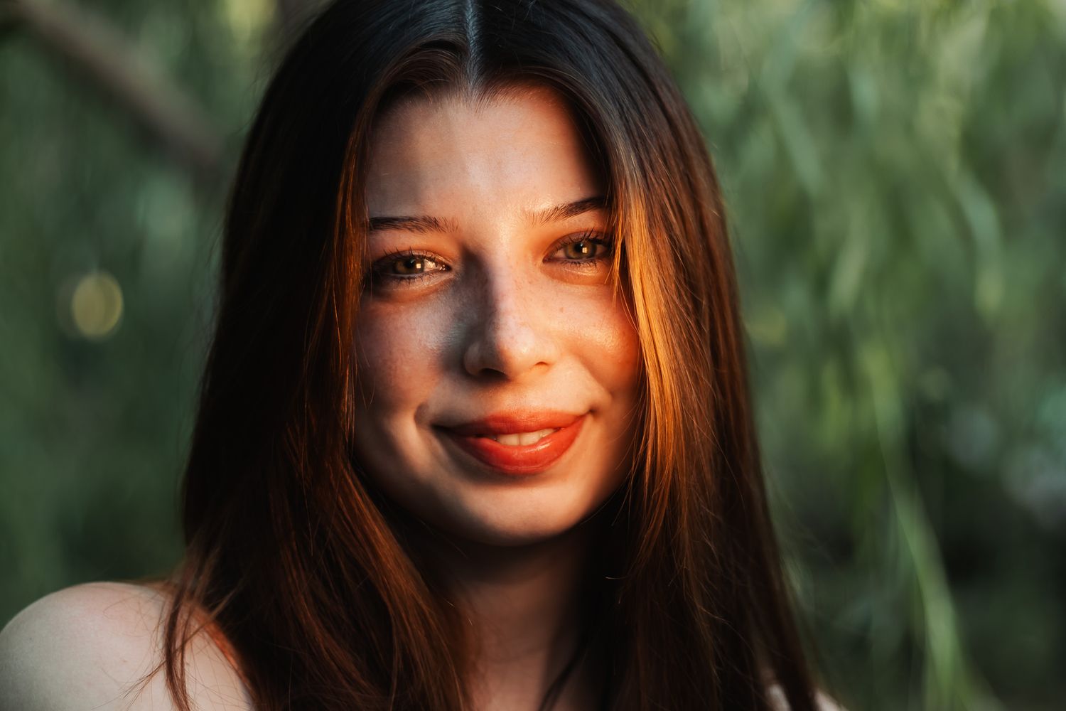 A smiling woman with long dark hair illuminated by warm sunlight against a soft green blurred background.