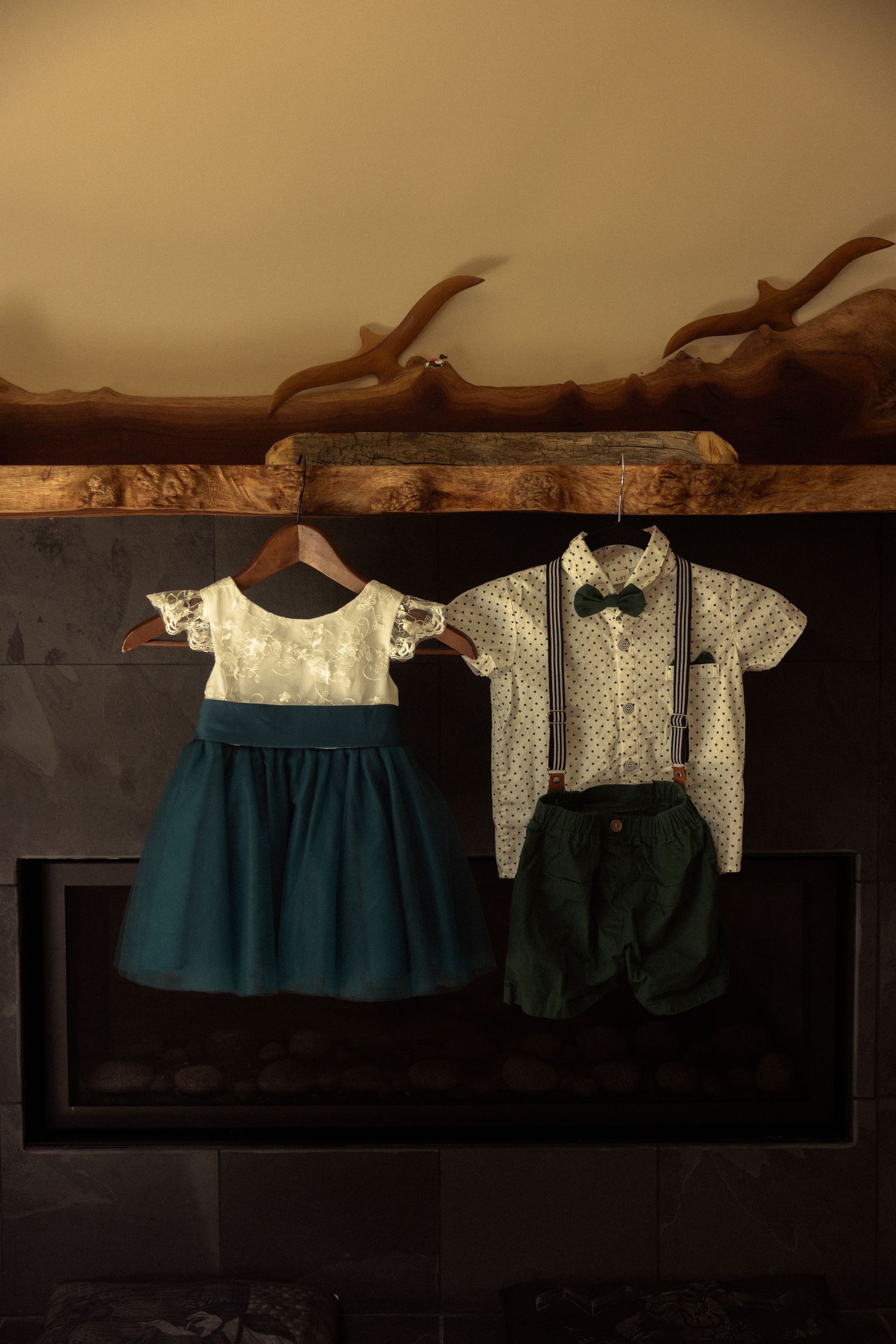Children's formal outfits hanging on wooden mantle, teal dress and boy's suspender shorts set with bow tie.