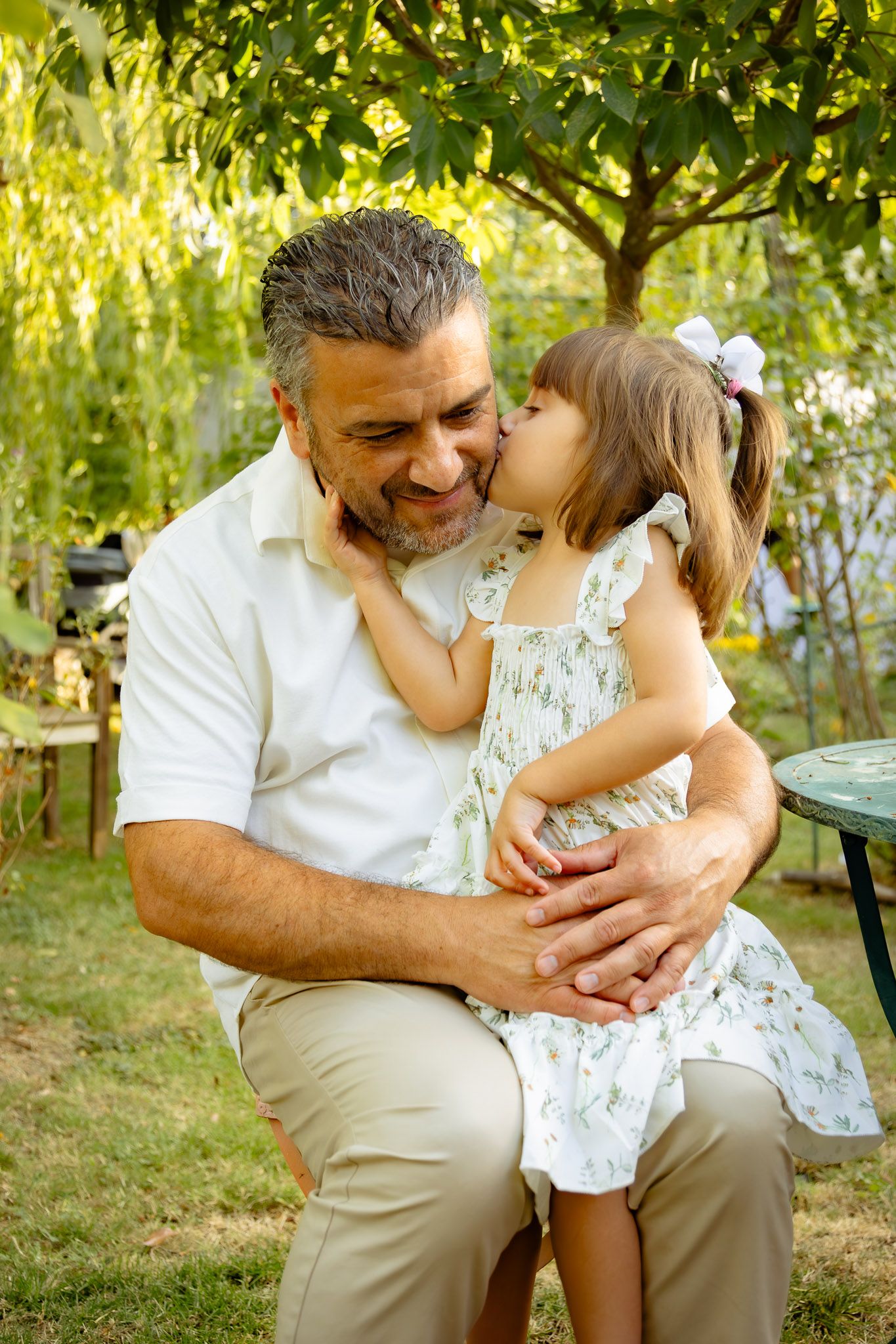 A sweet moment between a person in a white shirt and beige pants and a small child in a white dress outdoors.