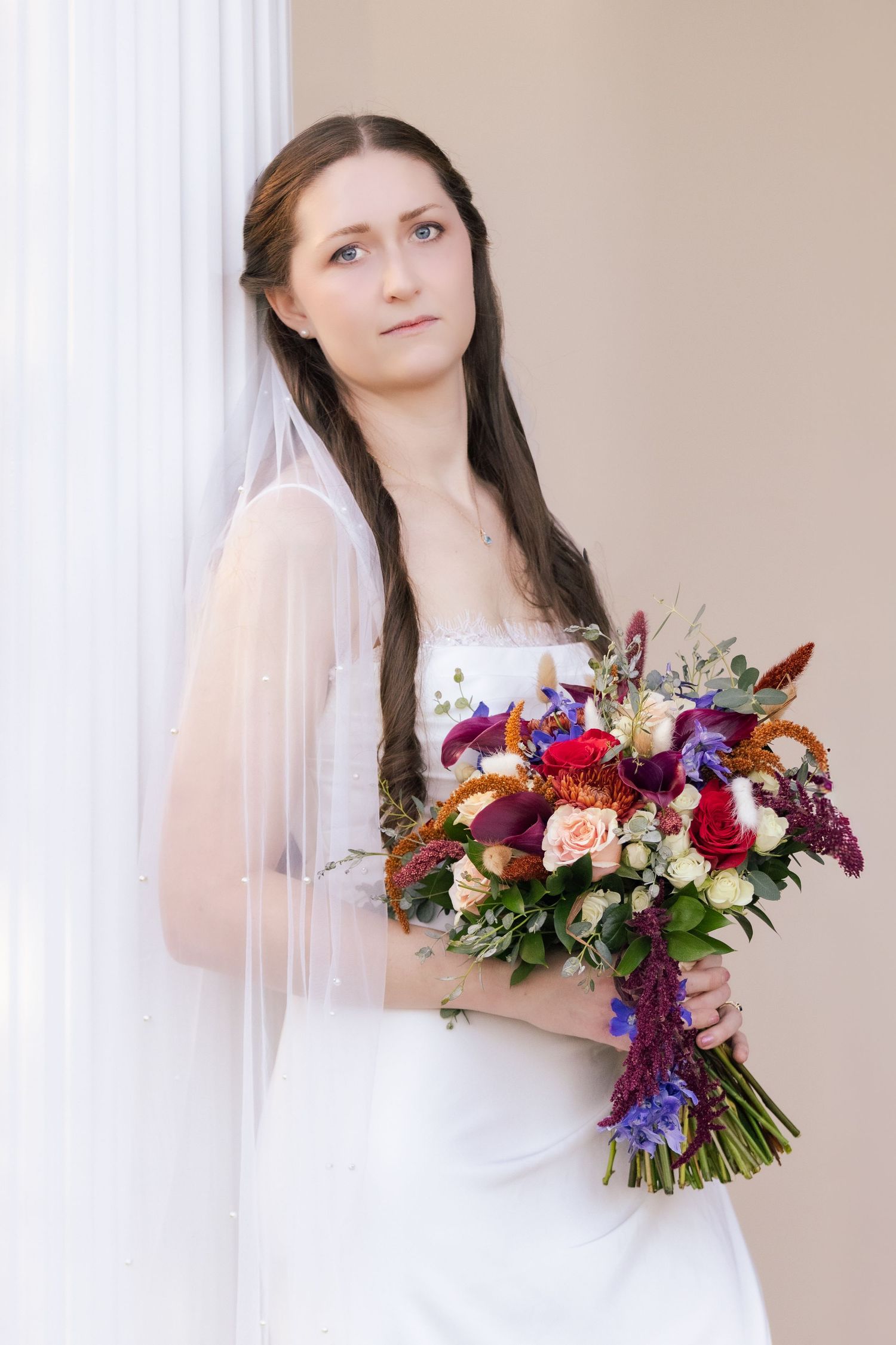 A bride in a white wedding dress holds a vibrant bouquet with red, purple and peach flowers against a neutral background.