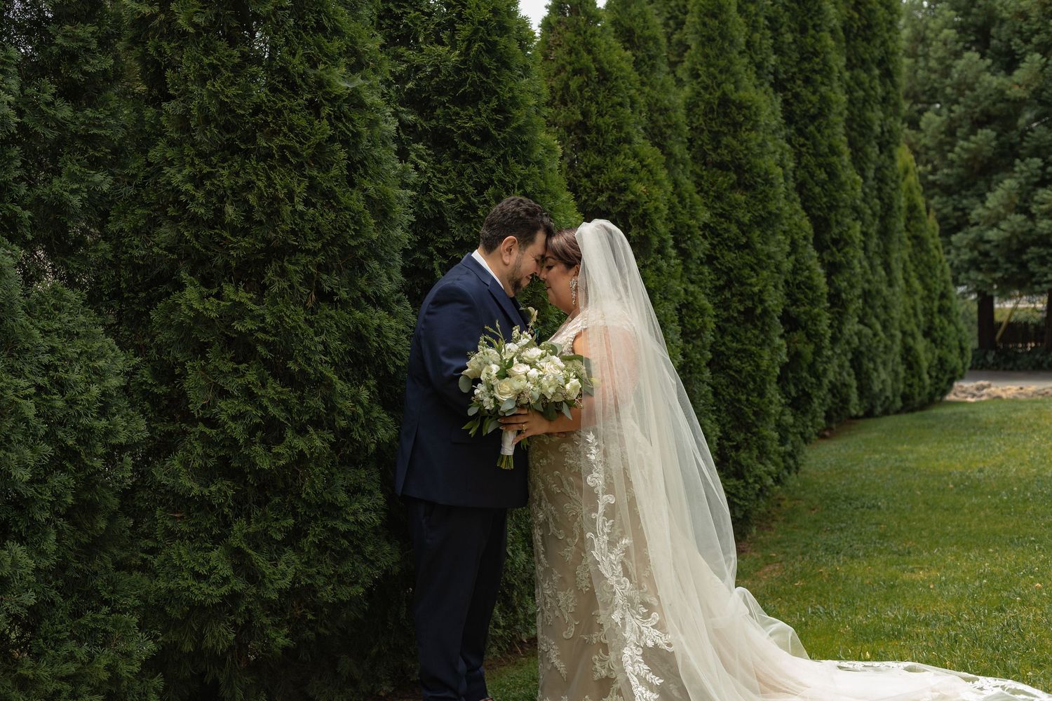 Bride and groom share a romantic moment by tall evergreen trees, bride holding white bouquet in lace gown with veil.