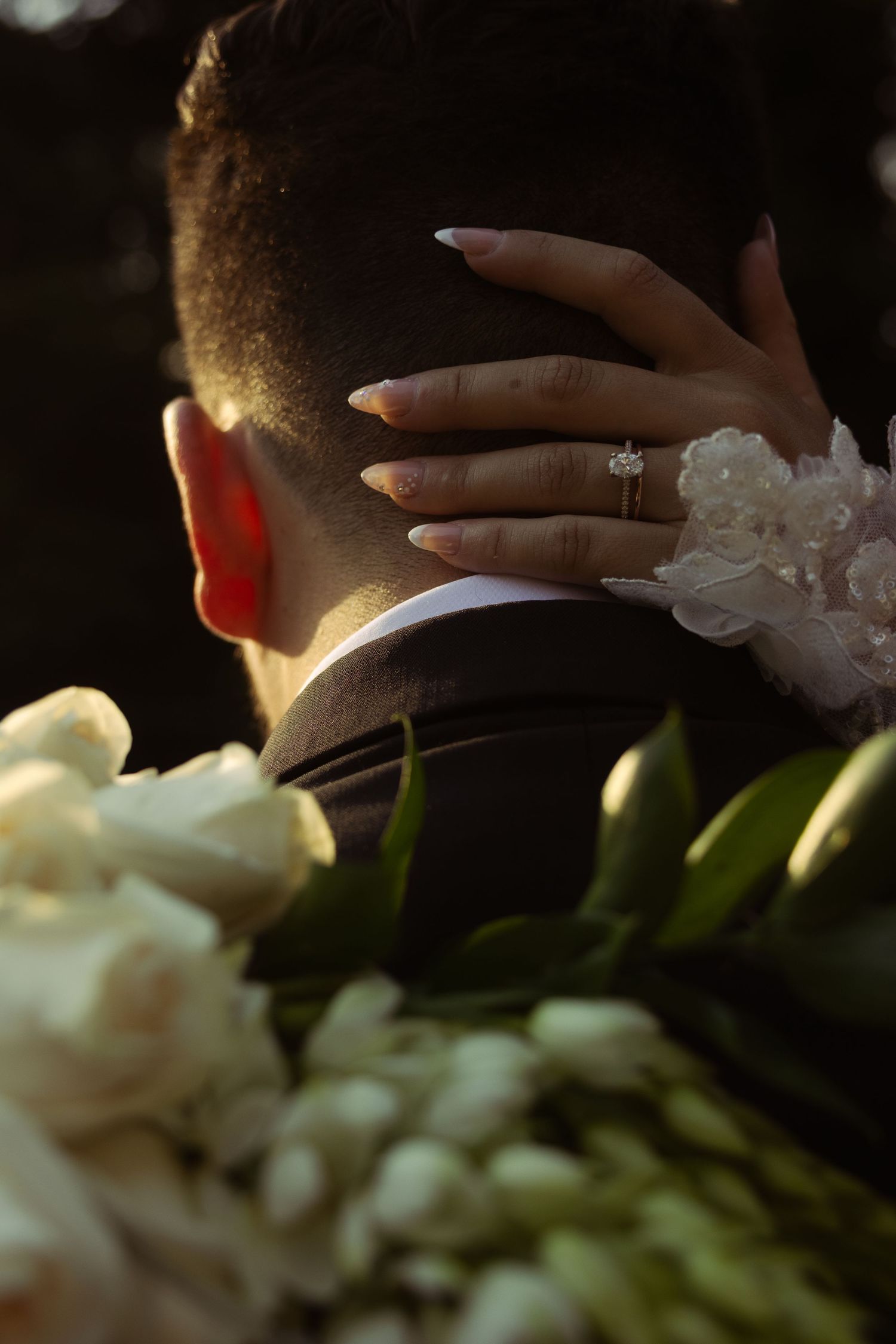 Bride with diamond engagement ring and lace sleeve rests hand on groom's neck, with white tulip bouquet in foreground.